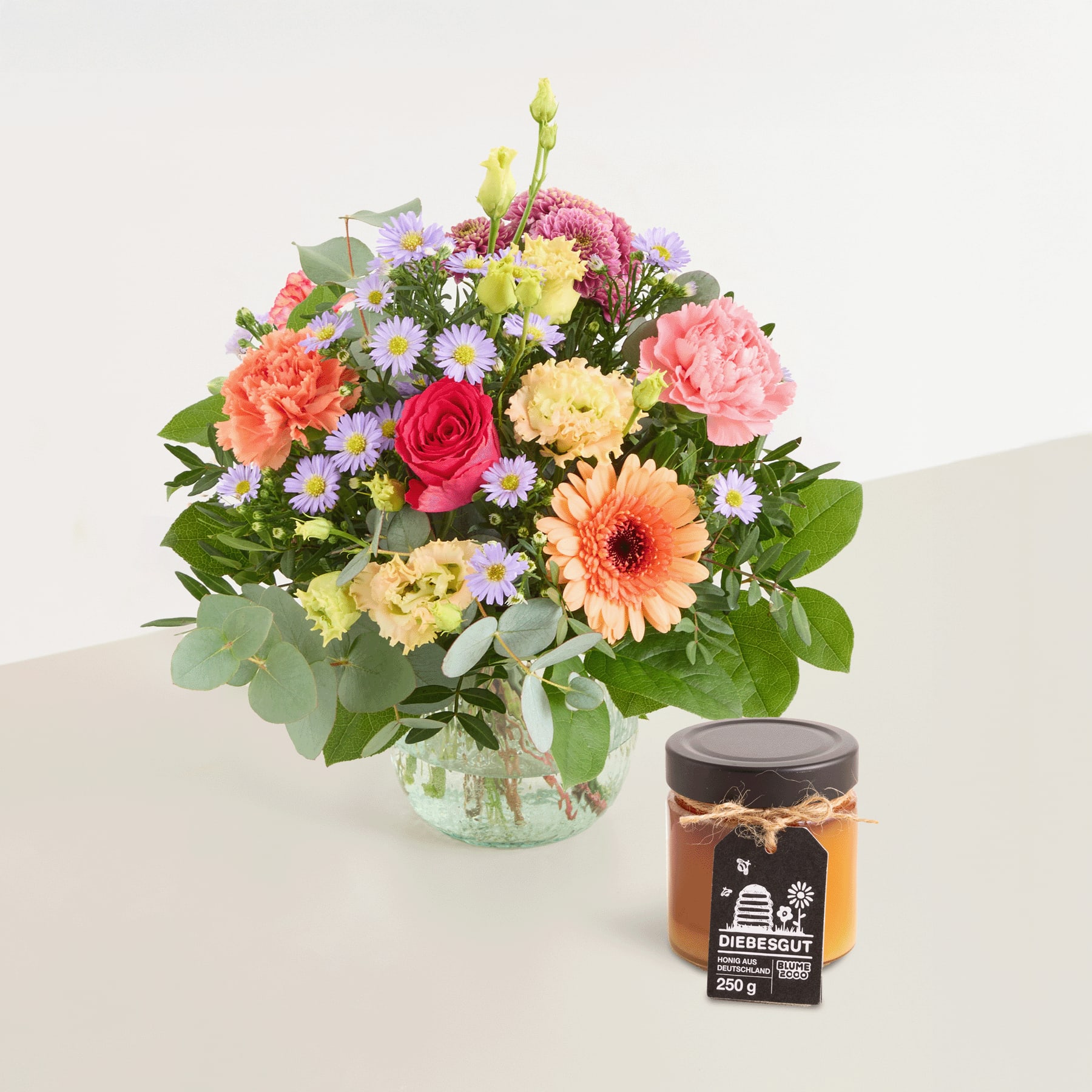 Ein leuchtender Strauß bunter Blumen, darunter Rosen, Gerbera und Chrysanthemen, steht in einer Glasvase auf einem Tisch, begleitet von einem Glas Honig.