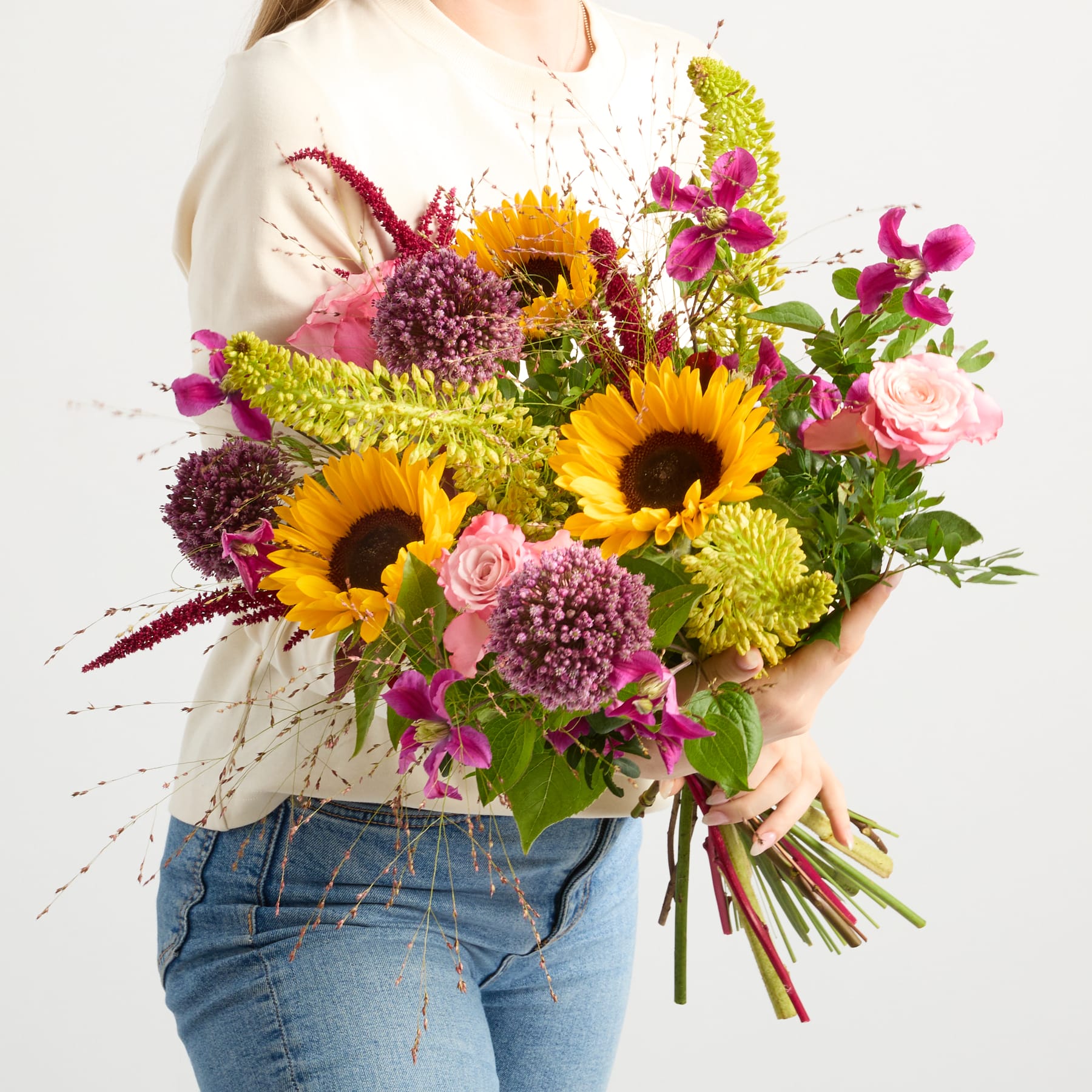 Ein farbenfroher Strauß aus Sonnenblumen, Rosen und anderen bunten Blumen wird von einer Person in einem weißen Hemd und Jeans vor einem schlichten Hintergrund gehalten.