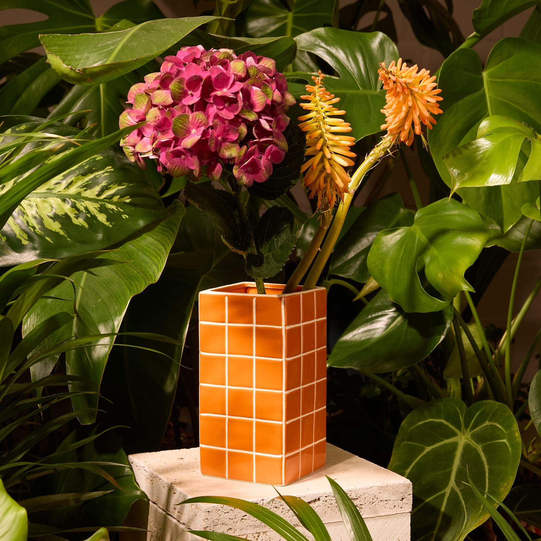 Eine leuchtend orangefarbene Keramikvase mit Gittermuster hält einen Strauß zarter rosa und gelber Blumen vor einem schlichten weißen Hintergrund.