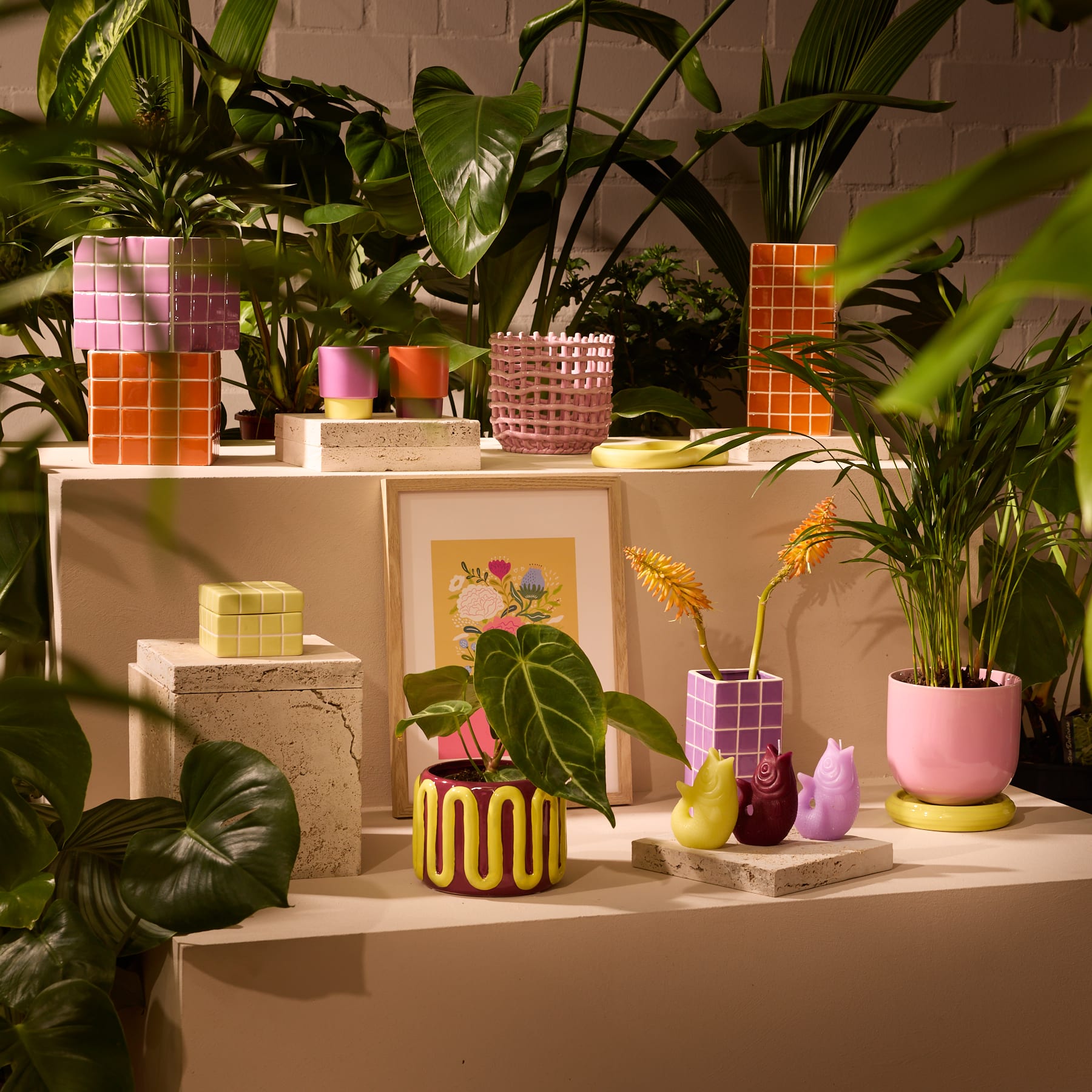 Eine lebendige und vielseitige Ausstellung farbenfroher Keramikvasen, Skulpturen und gerahmter Kunstwerke, umgeben von üppigem, grünem Laub vor einem neutralen Hintergrund.