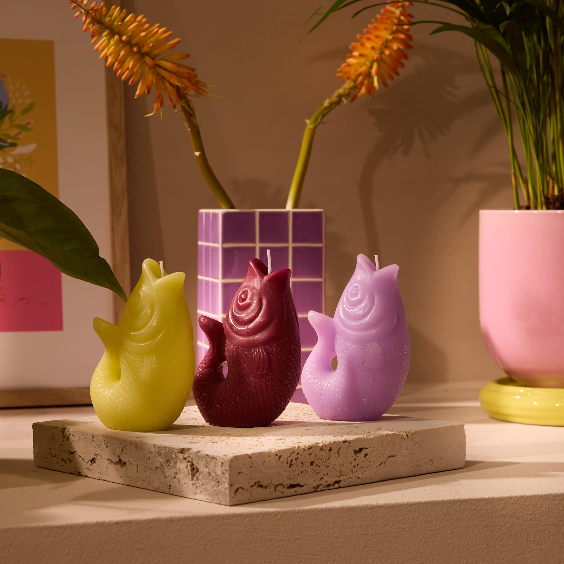 Das Bild zeigt eine Sammlung farbenfroher Keramikfiguren, darunter eine gelbe Ente, eine burgunderfarbene Rose und zwei violette Formen, die auf einer Betonplattform mit leuchtend orangefarbenen Blumen im Hintergrund angeordnet sind.