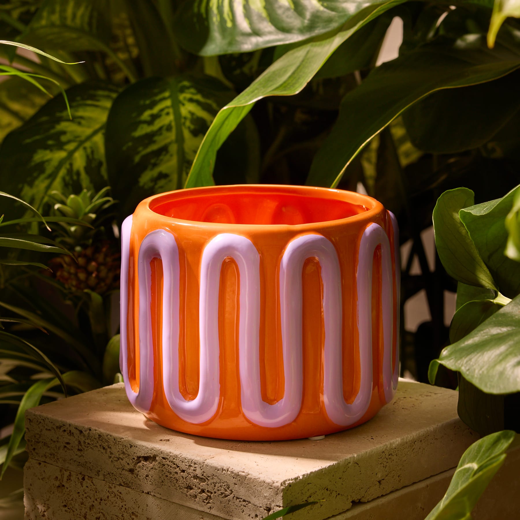Das Bild zeigt einen orangefarbenen Blumentopf mit einem wellenförmigen, abstrakten Muster in Rosa und Weiß, der eine grüne Blattpflanze enthält.