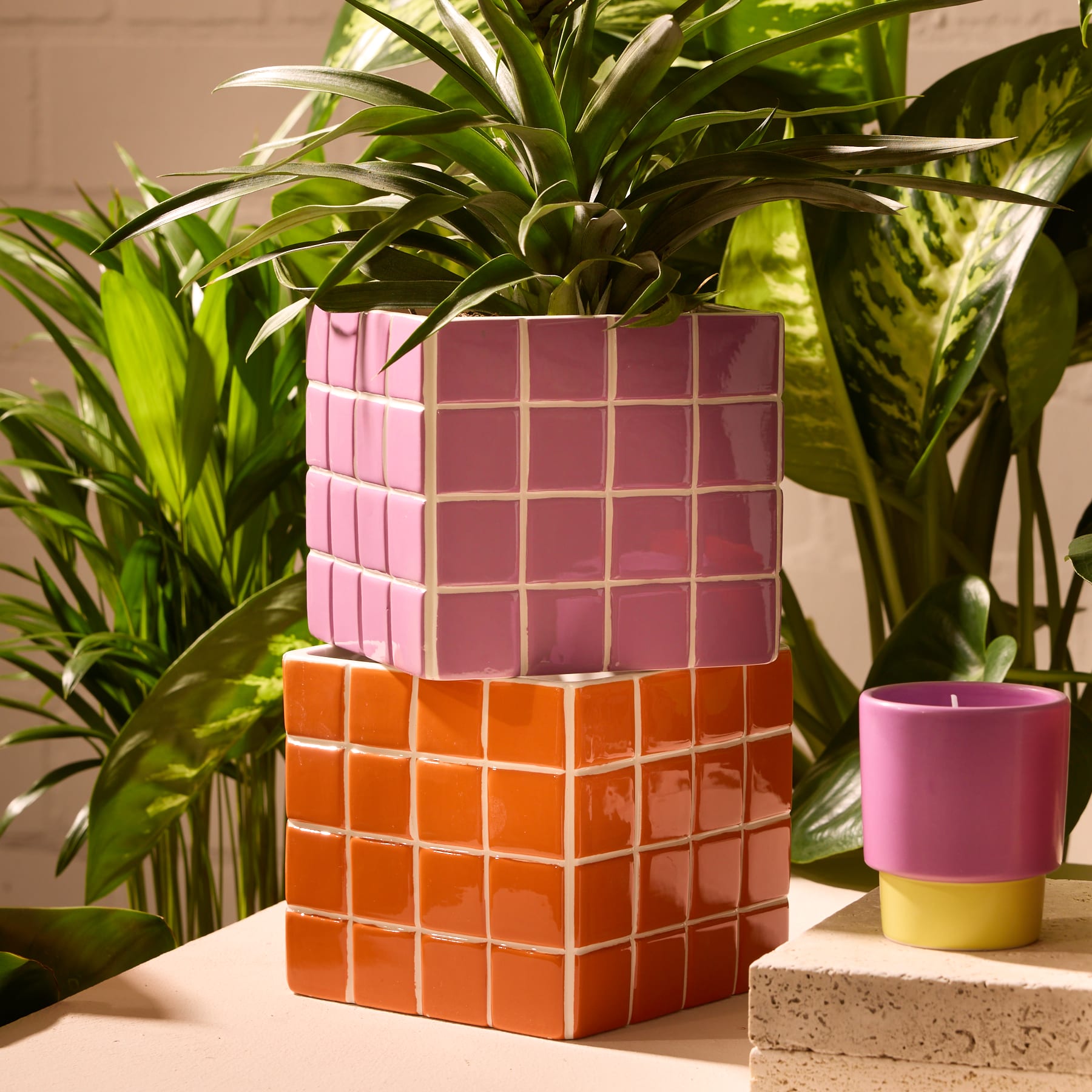 Das Bild zeigt einen Stapel farbenfroher, geometrischer Pflanzgefäße in verschiedenen Rosa- und Orangetönen, umgeben von üppigem, grünem Laub im Hintergrund.