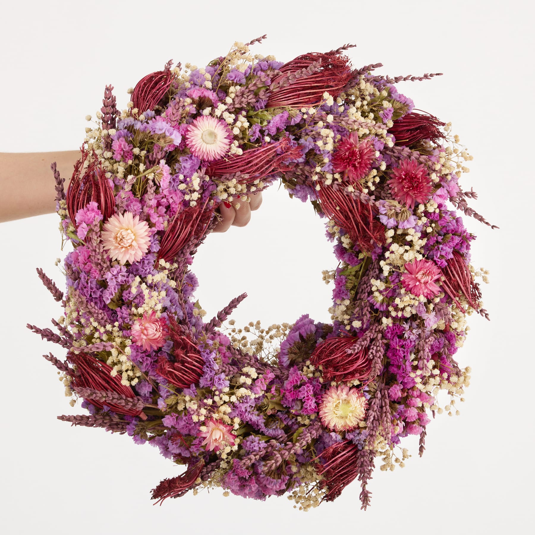 Ein leuchtender, runder Kranz aus verschiedenen violetten und weißen Blumen, den eine Person vor einem schlichten Hintergrund in den Händen hält.