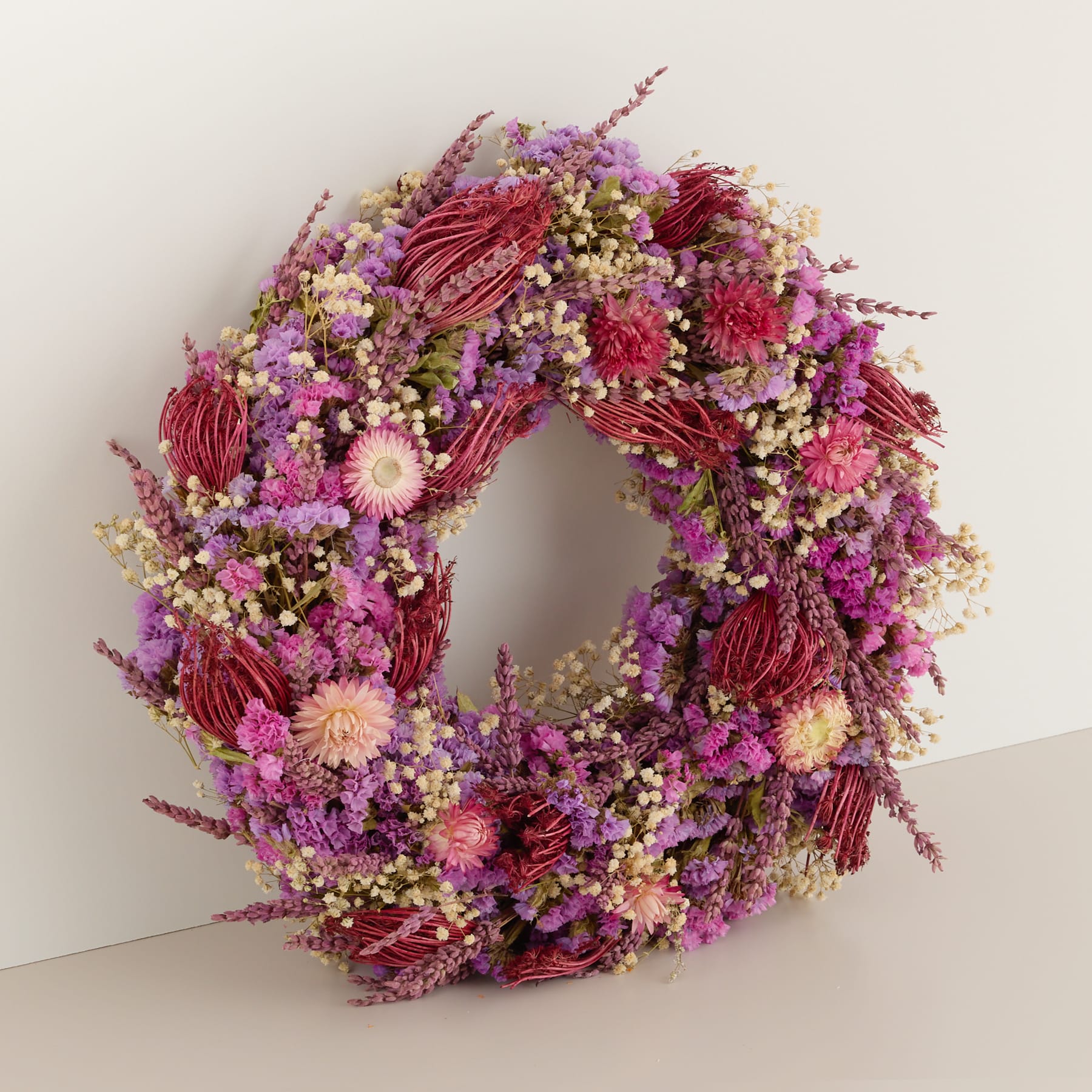 Ein lebendiger und farbenfroher Kranz aus verschiedenen getrockneten Blumen, darunter in Rot- und Lilatönen, schmückt eine schlichte weiße Wand.