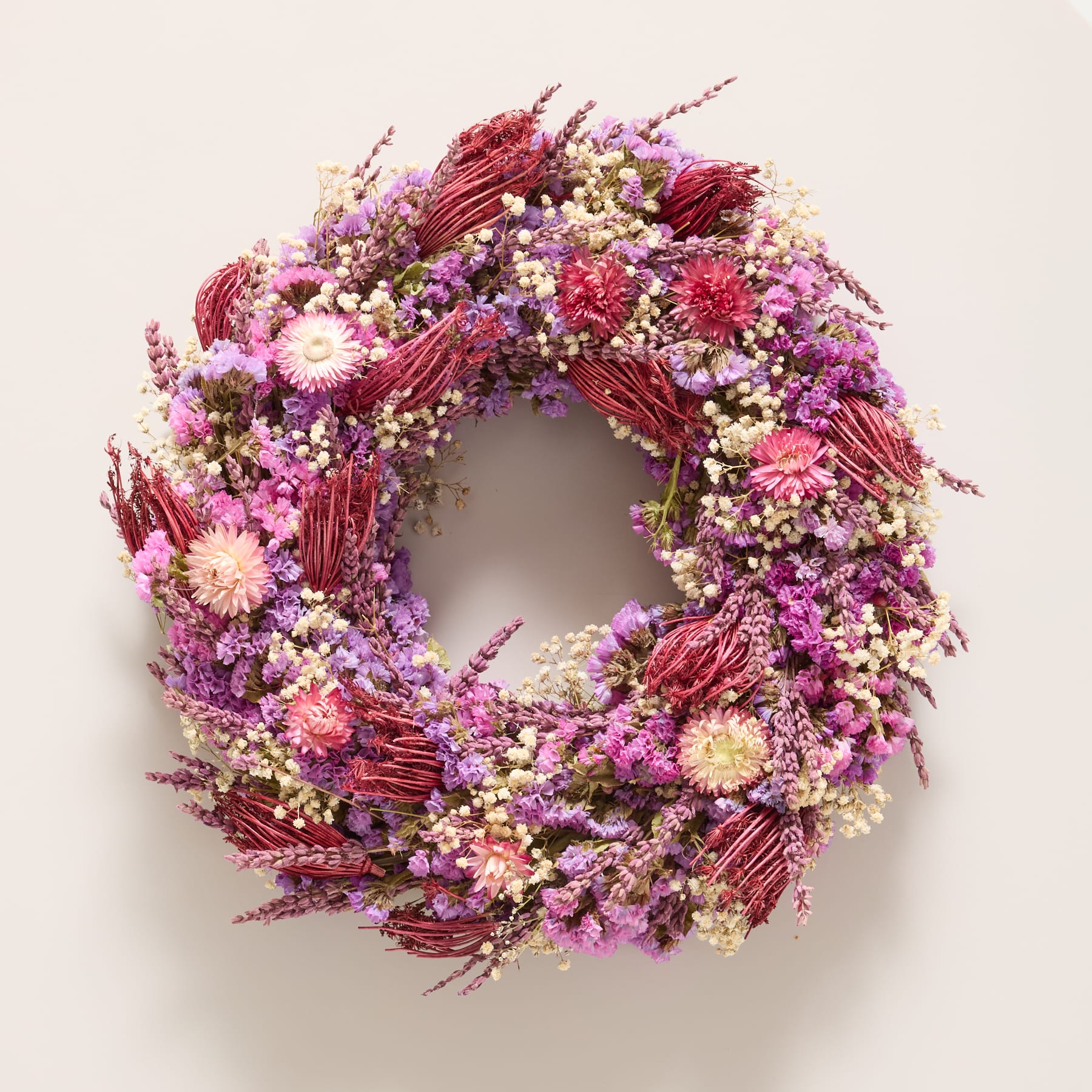 Ein lebendiger und farbenfroher Kranz aus verschiedenen getrockneten Blumen in Rot-, Rosa- und Lilatönen ziert den neutral getönten Hintergrund.