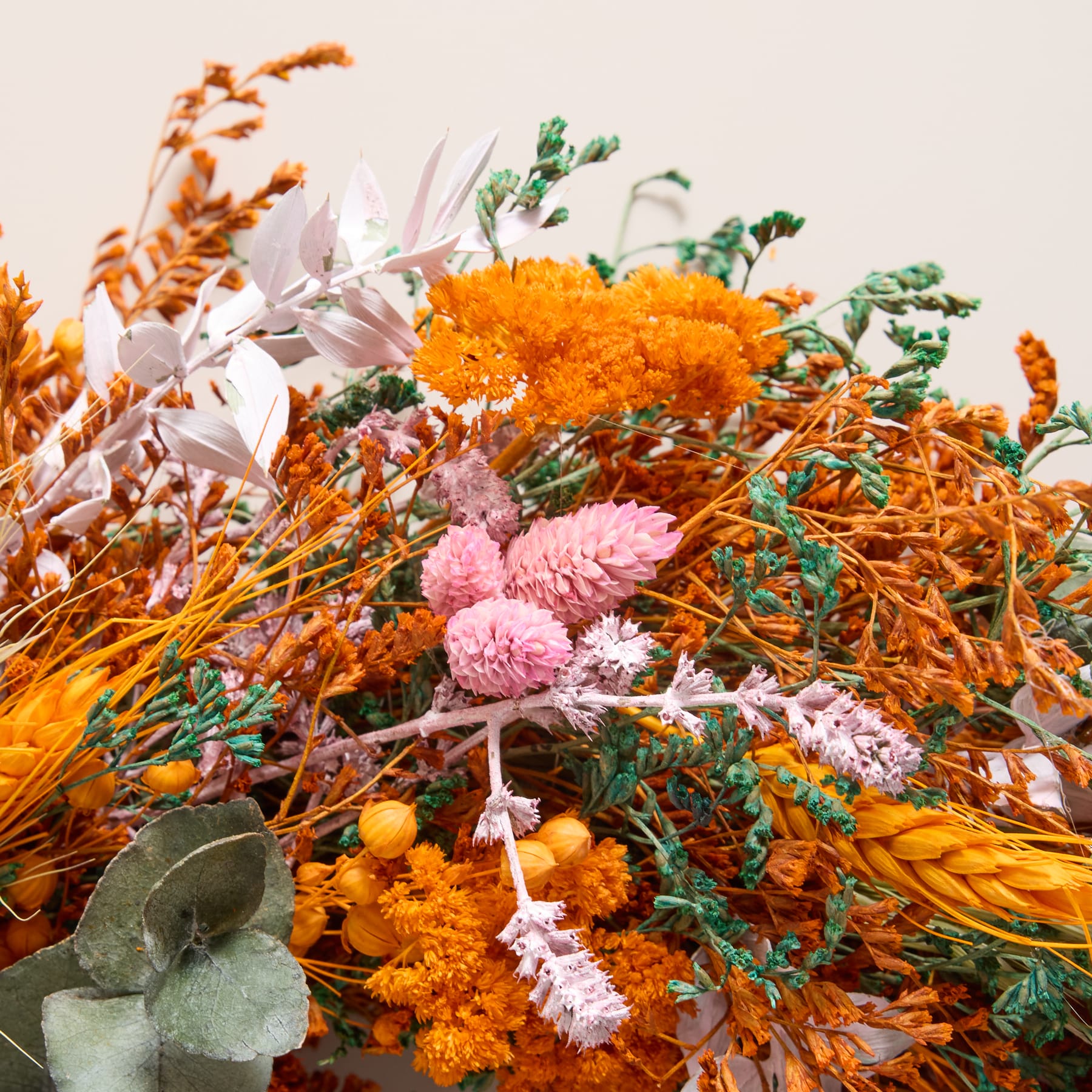 Ein farbenfroher, herbstlicher Kranz aus getrockneten Blumen, Blättern und Gräsern wird vor einem schlichten weißen Hintergrund in einer Hand gehalten.