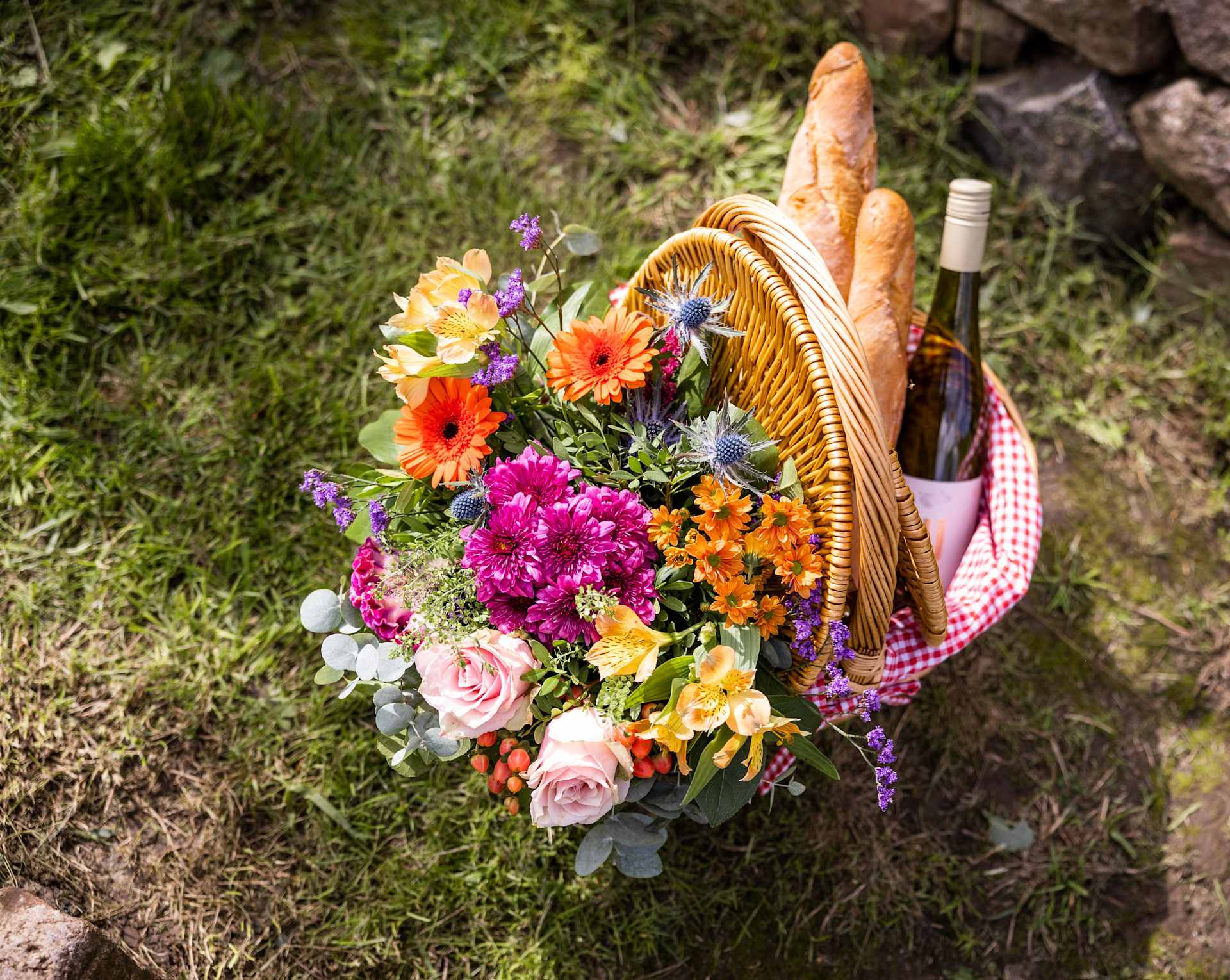 Ein farbenfroher Korb mit einer bunten Auswahl an Blumen, darunter Rosen, Gänseblümchen und andere Blüten, steht vor einer Kulisse aus üppigem Grün und felsigem Gelände.