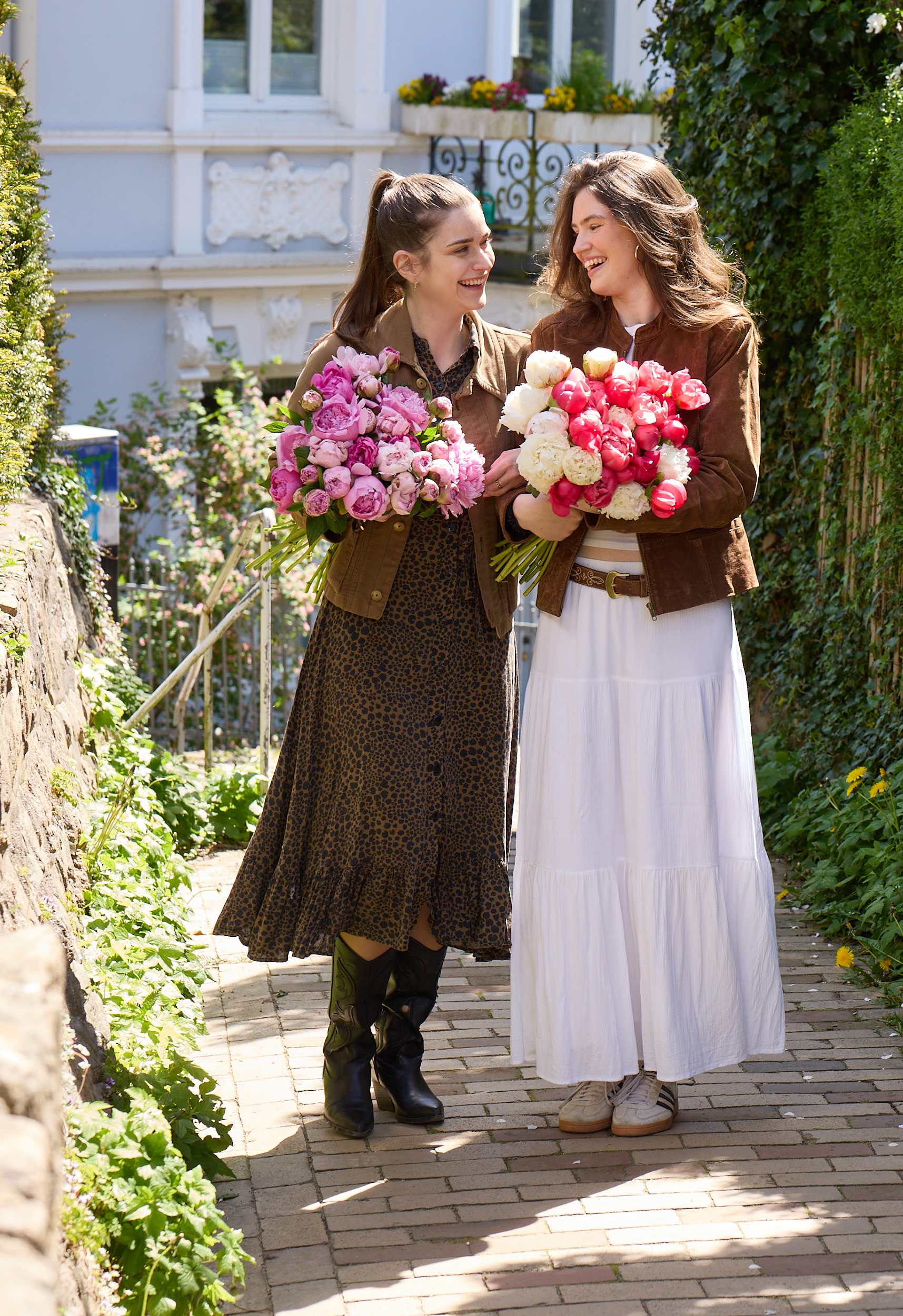 Zwei Frauen in Freizeitkleidung gehen zusammen spazieren und tragen große Sträuße leuchtend rosa Blumen vor der Kulisse einer bezaubernden Gartenanlage mit üppigem Grün und einem Steinweg.