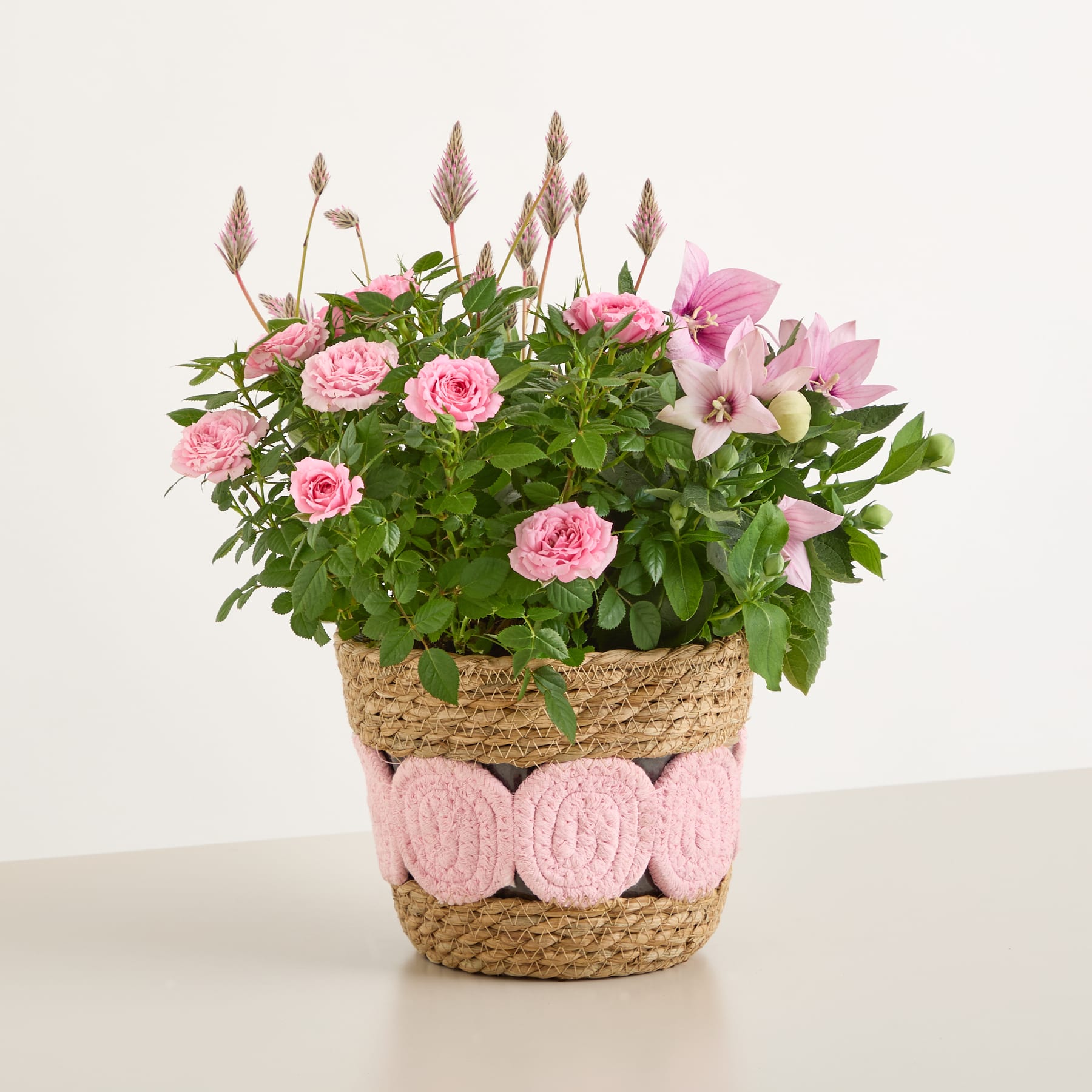 Ein farbenfrohes Arrangement aus rosa Rosen, Lilien und anderen Blumen in einem geflochtenen Korb vor einem schlichten weißen Hintergrund.