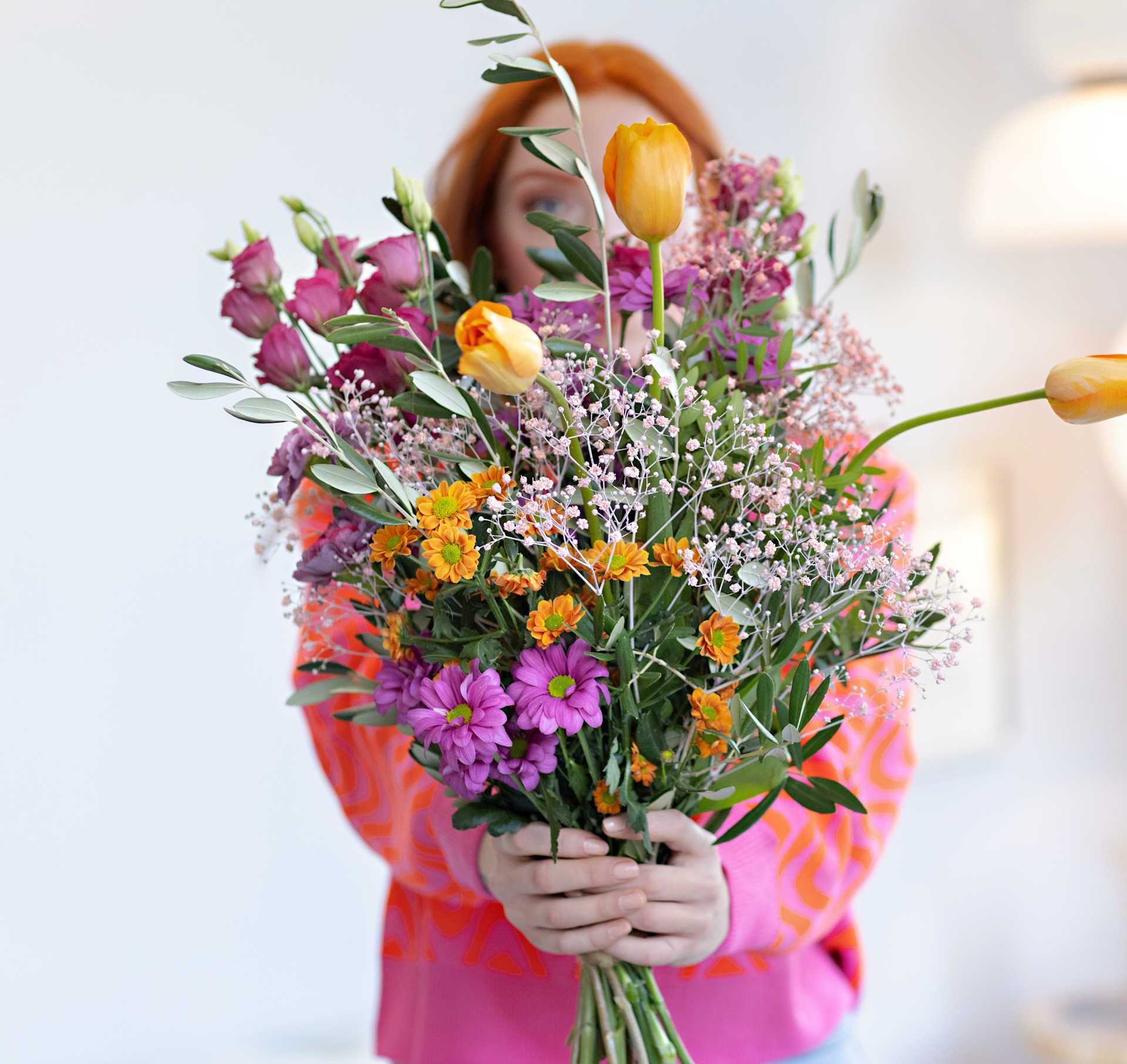Ein lebendiger Strauß bunter Blumen, darunter Tulpen, Gänseblümchen und andere Blüten, wird im Vordergrund vor einem unscharfen Hintergrund gehalten.