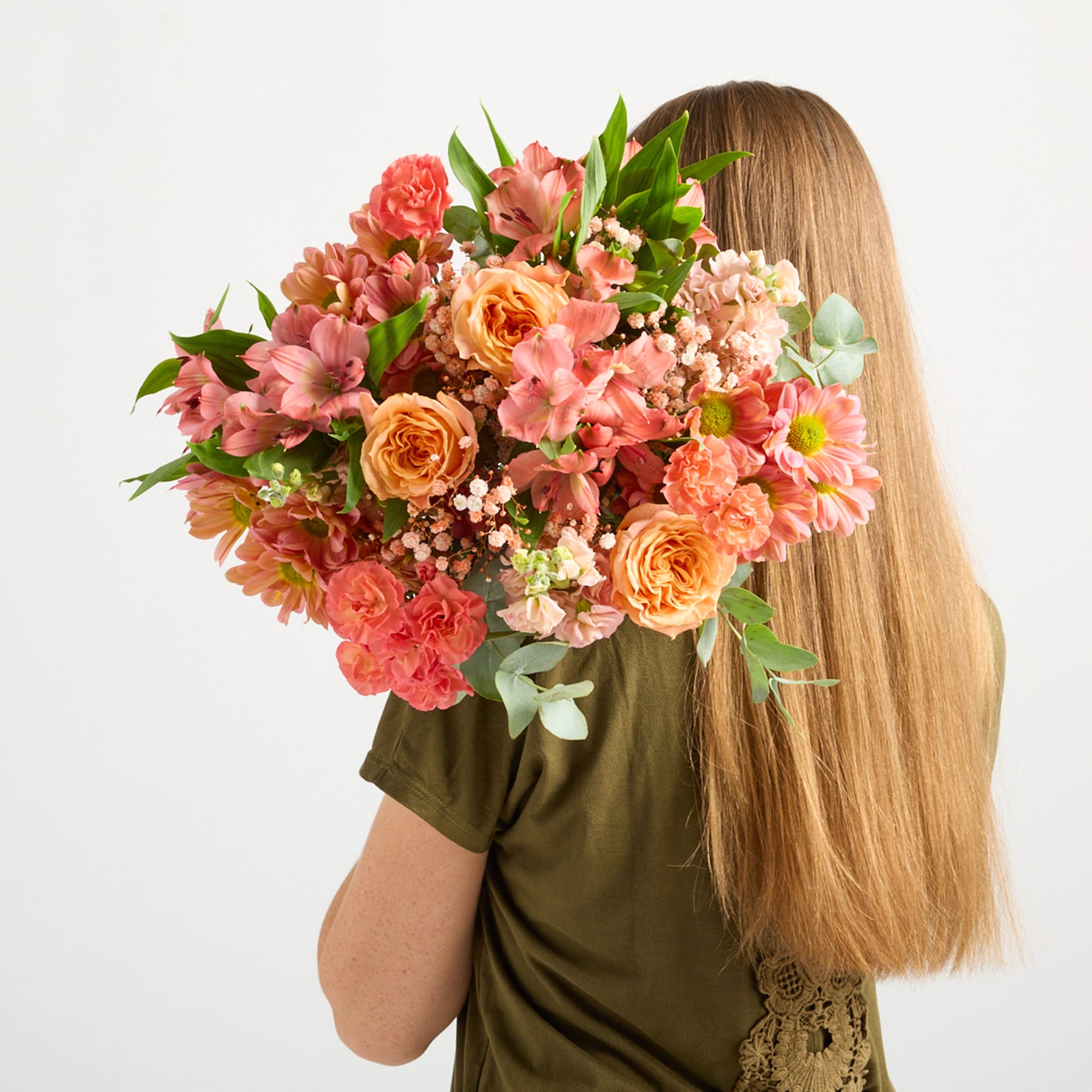 Ein leuchtender Strauß bunter Blumen, darunter Rosen, Nelken und Gänseblümchen, wird vor einem schlichten weißen Hintergrund vor eine Person mit langem, blondem Haar gehalten.