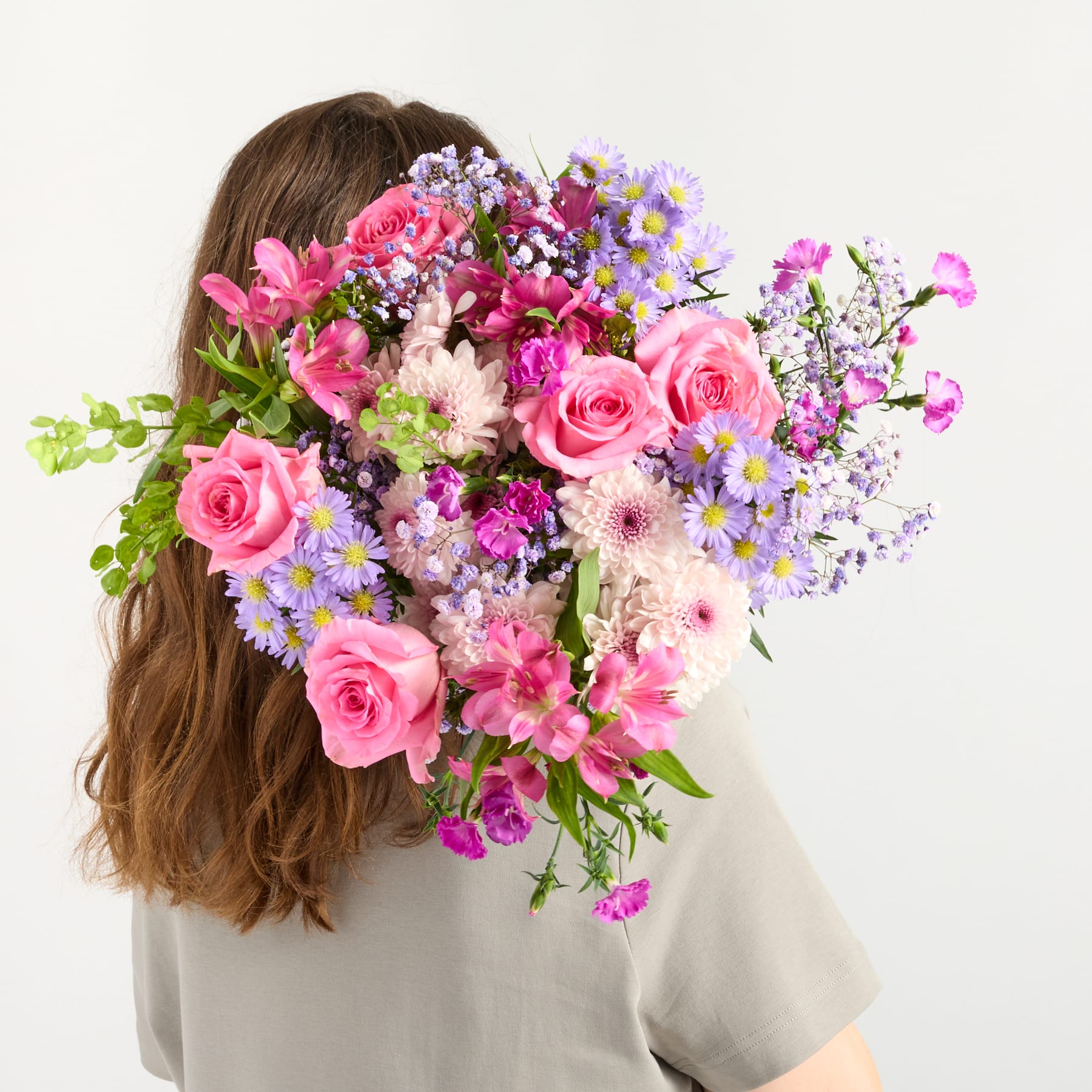Ein leuchtender Strauß aus rosa Rosen, lila Gänseblümchen und anderen bunten Blumen bedeckt das Gesicht einer Person mit langem, welligem braunem Haar vor einem schlichten weißen Hintergrund.