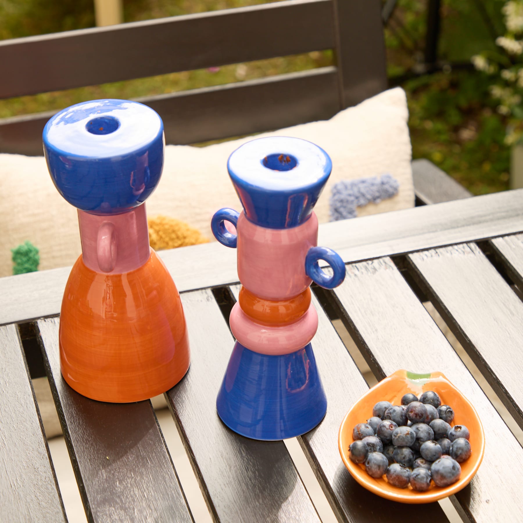 Zwei bunte Salz- und Pfefferstreuer aus Keramik stehen auf einer Holzbank. Im Vordergrund steht eine kleine Schale mit frischen Blaubeeren. Im Hintergrund ist eine üppige, grüne Außenlandschaft zu sehen.