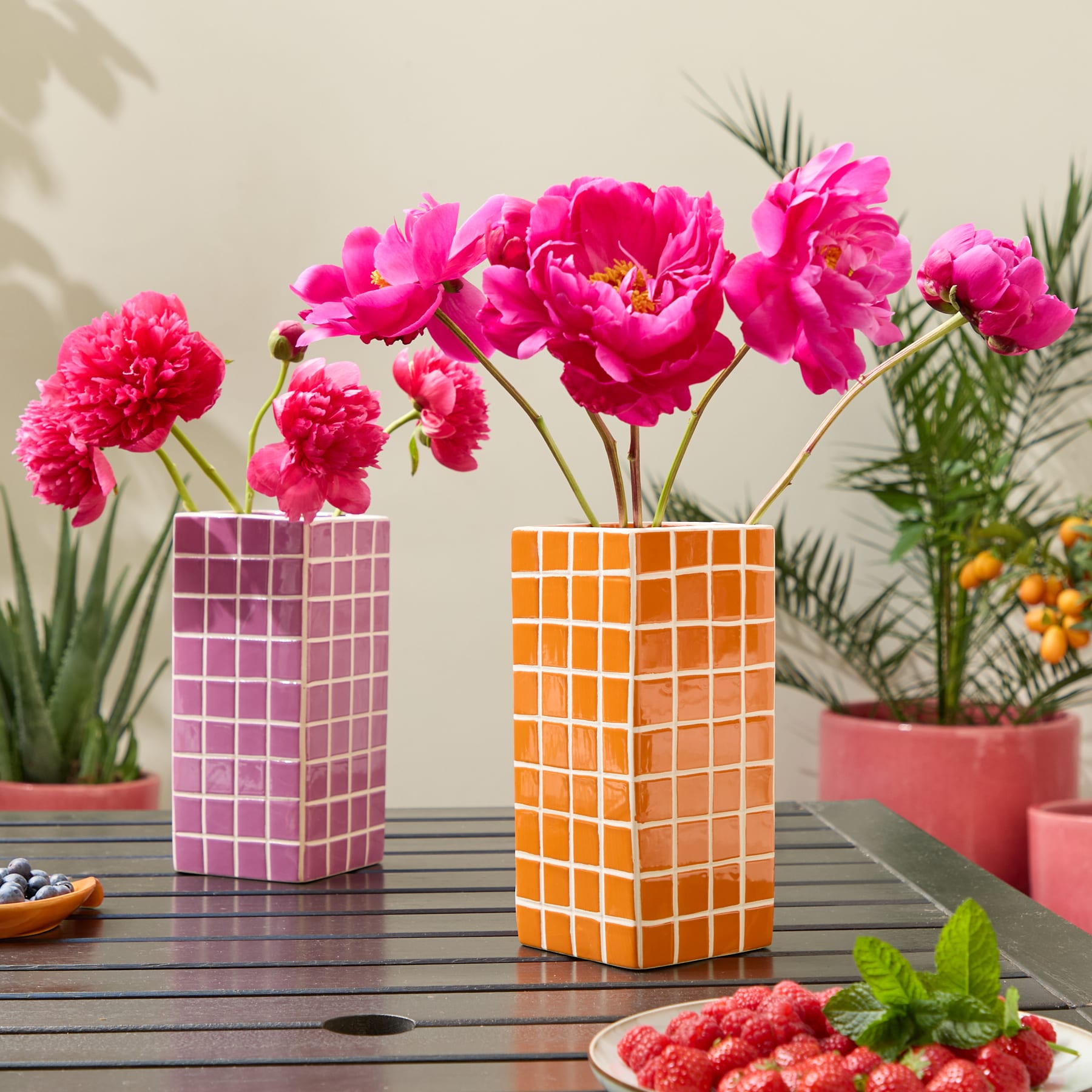 Leuchtend rosa Nelken in geometrischen Vasen stehen auf einem Holztisch, umgeben von Topfpflanzen vor einem neutralen Hintergrund.