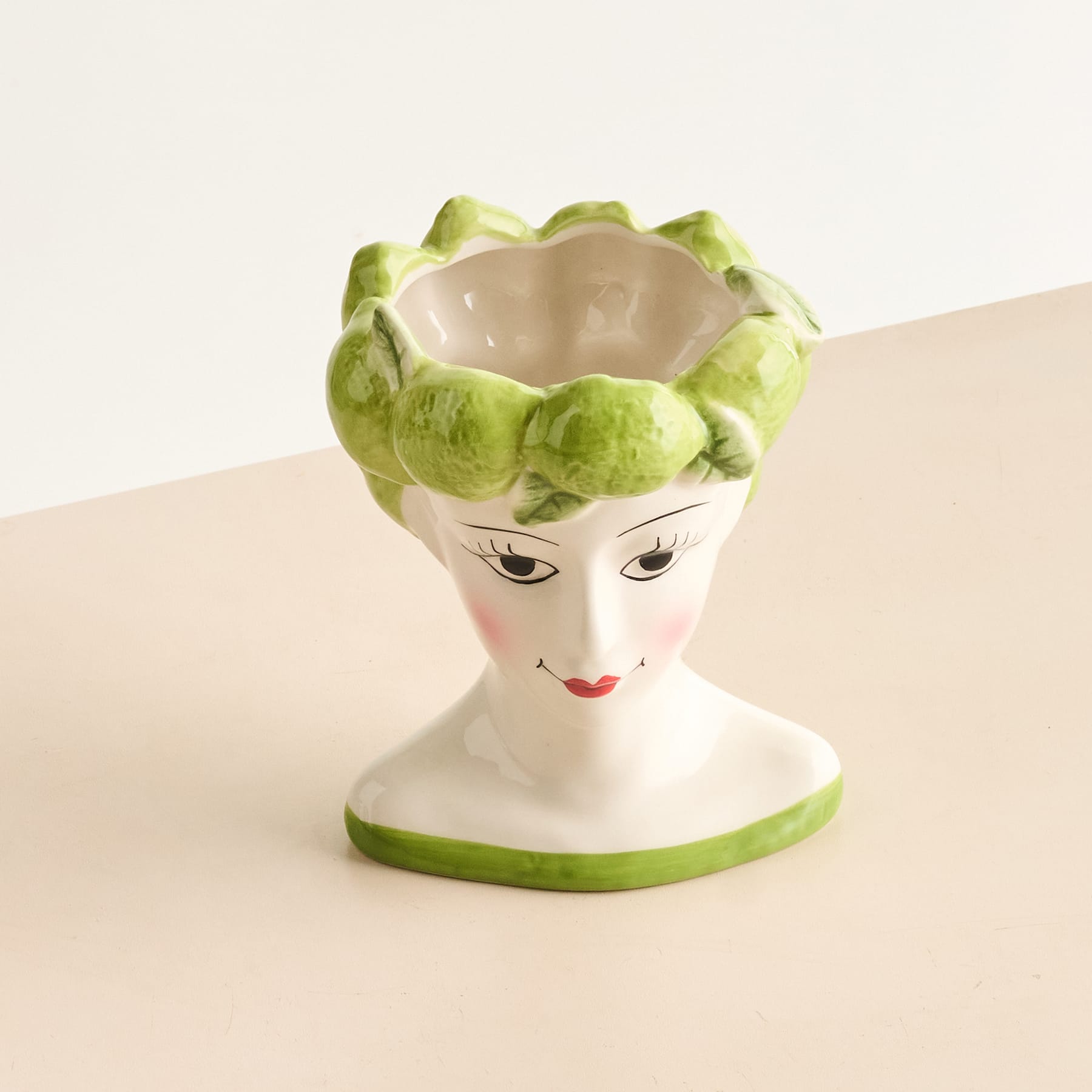 Eine Keramikfigur mit weißem Gesicht und grüner Blumenkrone sitzt auf einer beigen Oberfläche vor einem schlichten Hintergrund.