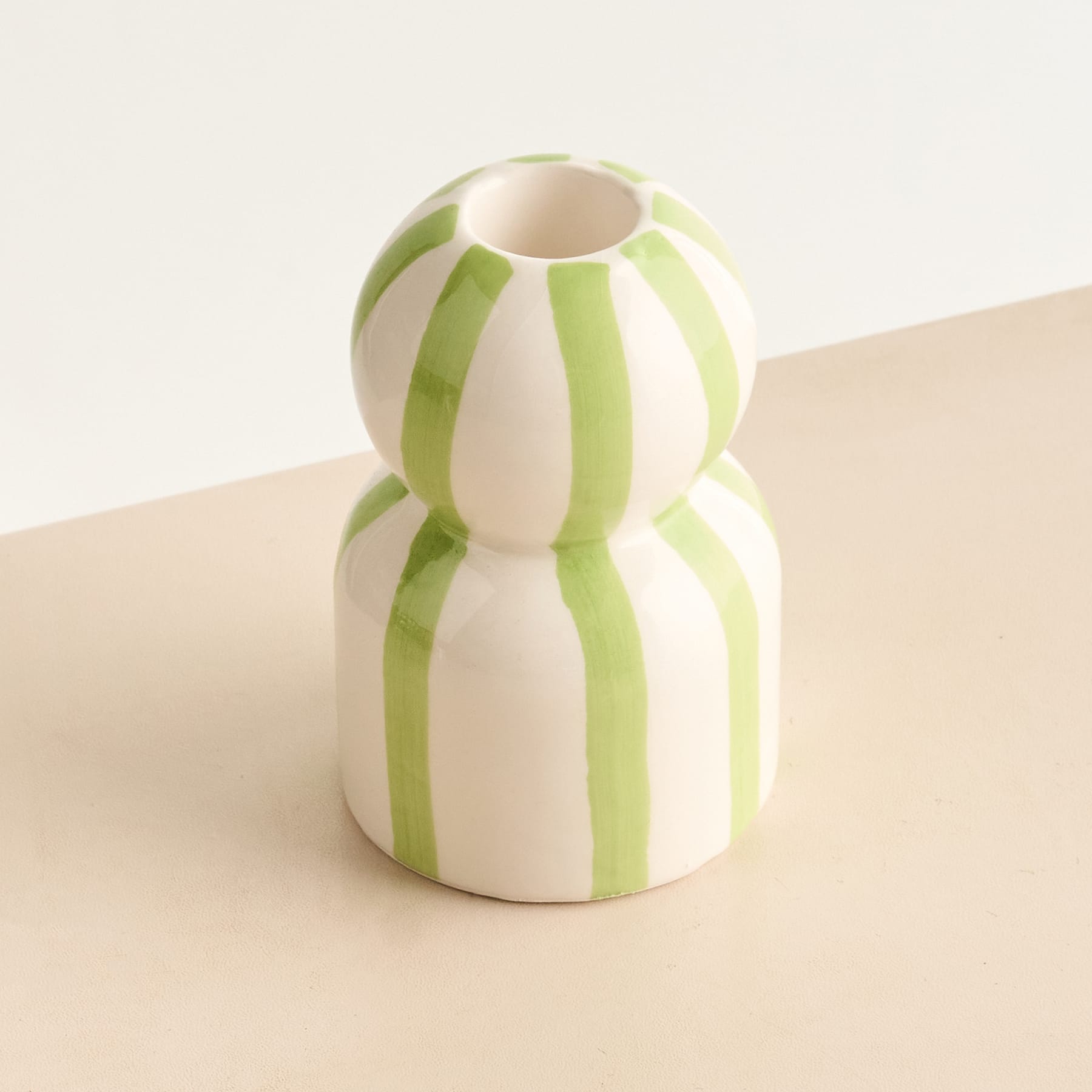 Eine weiß-grün gestreifte Keramikvase oder ein Kerzenhalter steht auf einer beigen Oberfläche vor einem schlichten Hintergrund.