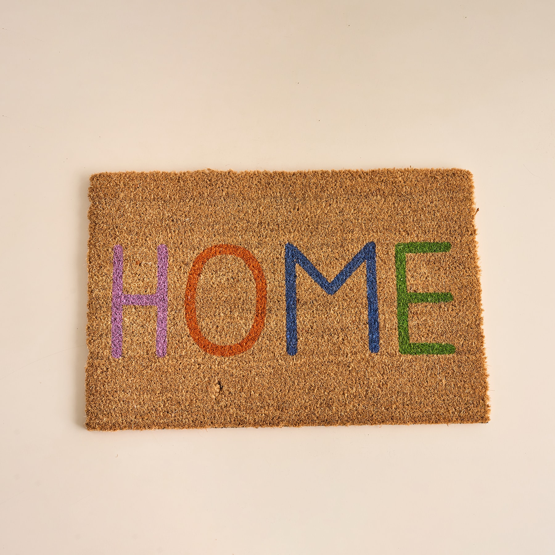 Auf einem schlichten beigen Untergrund liegt eine farbenfrohe „HOME“-Fußmatte aus gewebten Naturfasern.