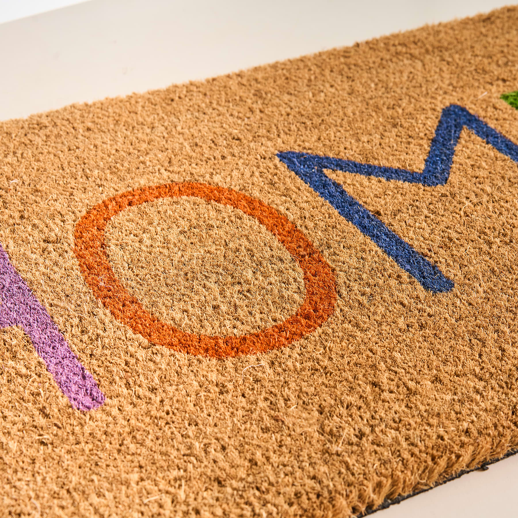 Das Bild zeigt eine Fußmatte mit dem Wort „OM“ in orangefarbenen und blauen Buchstaben auf beigem Hintergrund.