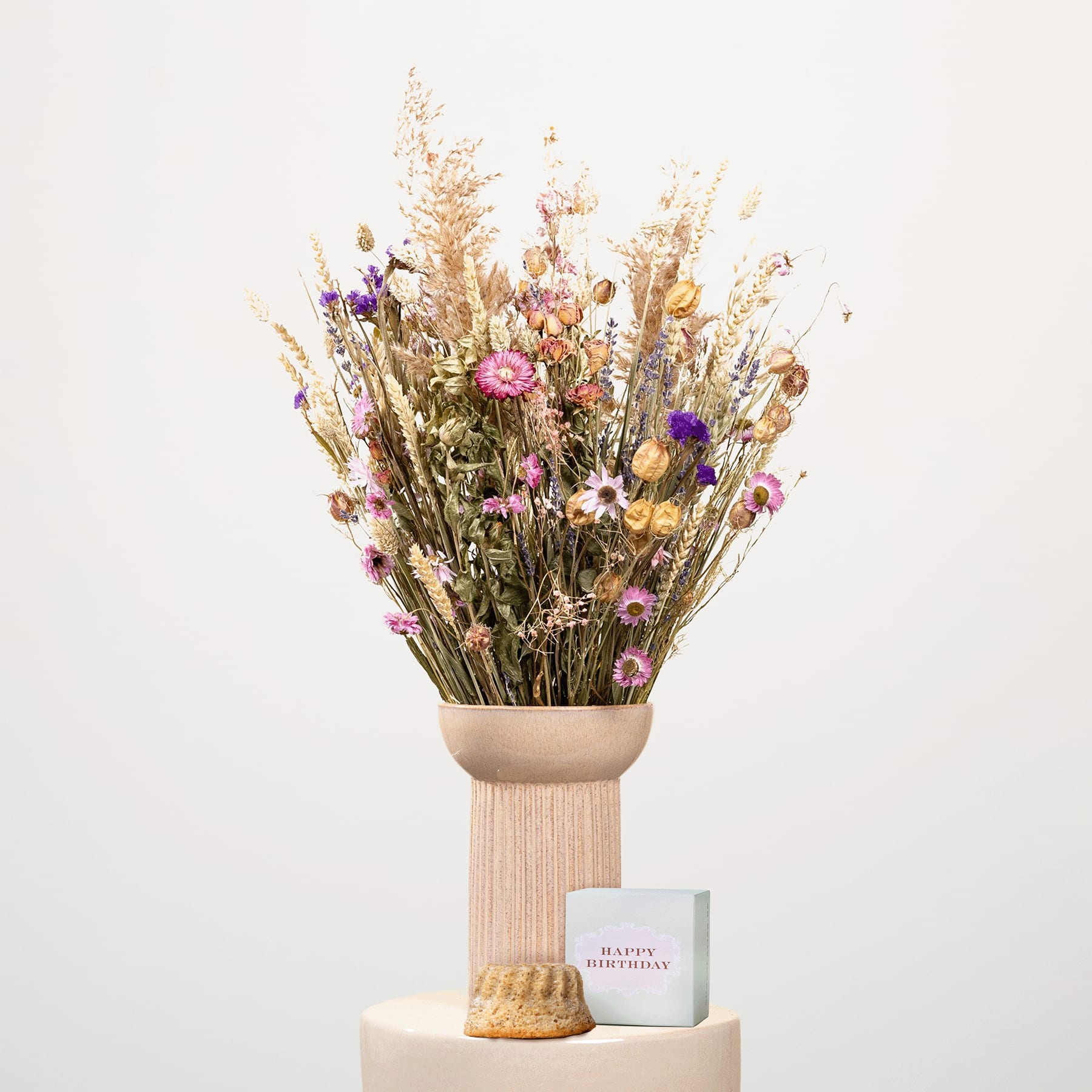 Ein Strauß leuchtender Trockenblumen, darunter rosa und violette Blüten, steht auf einem Holzsockel vor einem schlichten weißen Hintergrund.