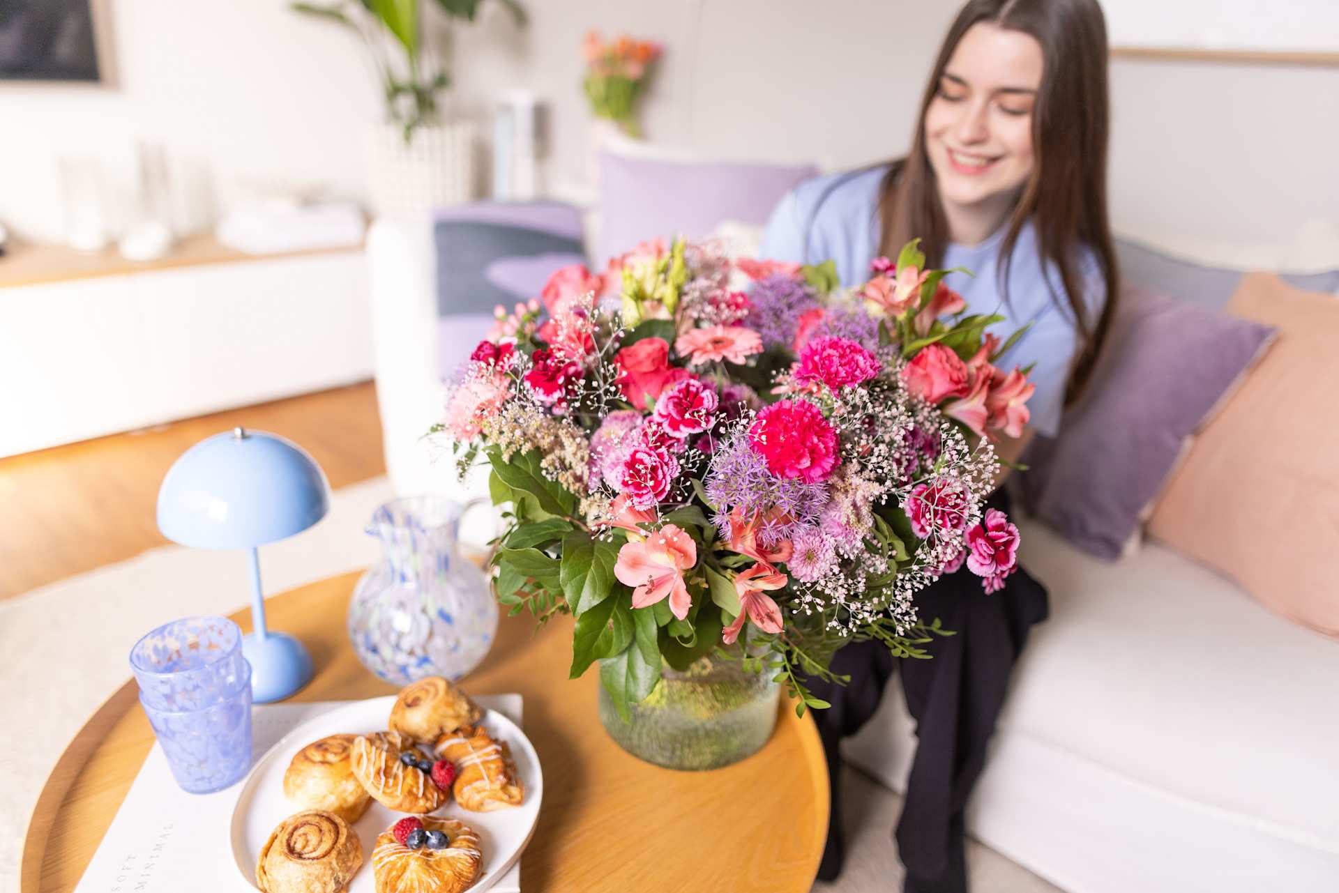 Auf einem Holztisch steht ein farbenfroher Strauß bunter Blumen, im Hintergrund ist eine lächelnde junge Frau zu sehen.