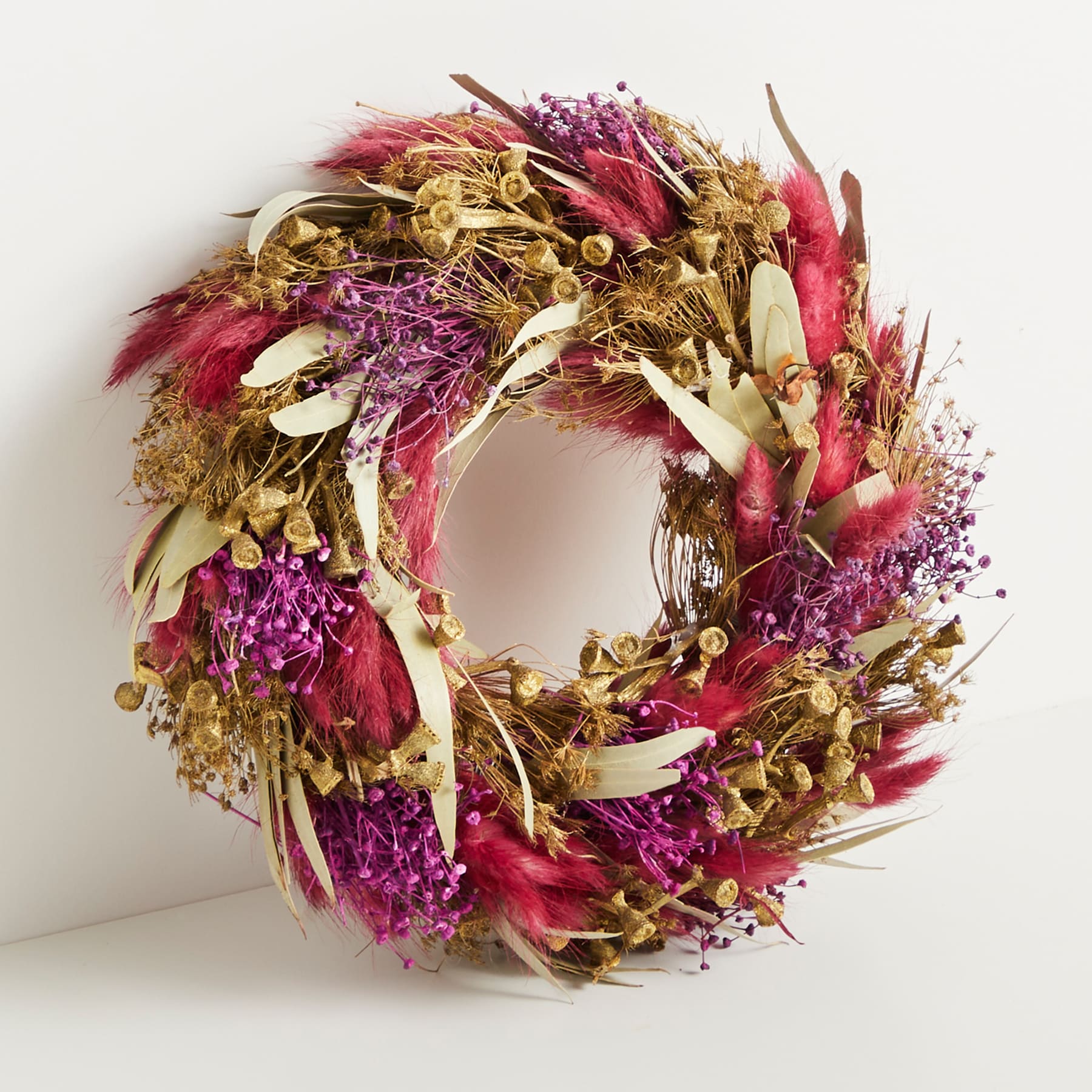 Ein leuchtender, getrockneter Blumenkranz aus Blumen und Blättern in verschiedenen Rosa-, Lila- und Goldtönen vor einem schlichten weißen Hintergrund.