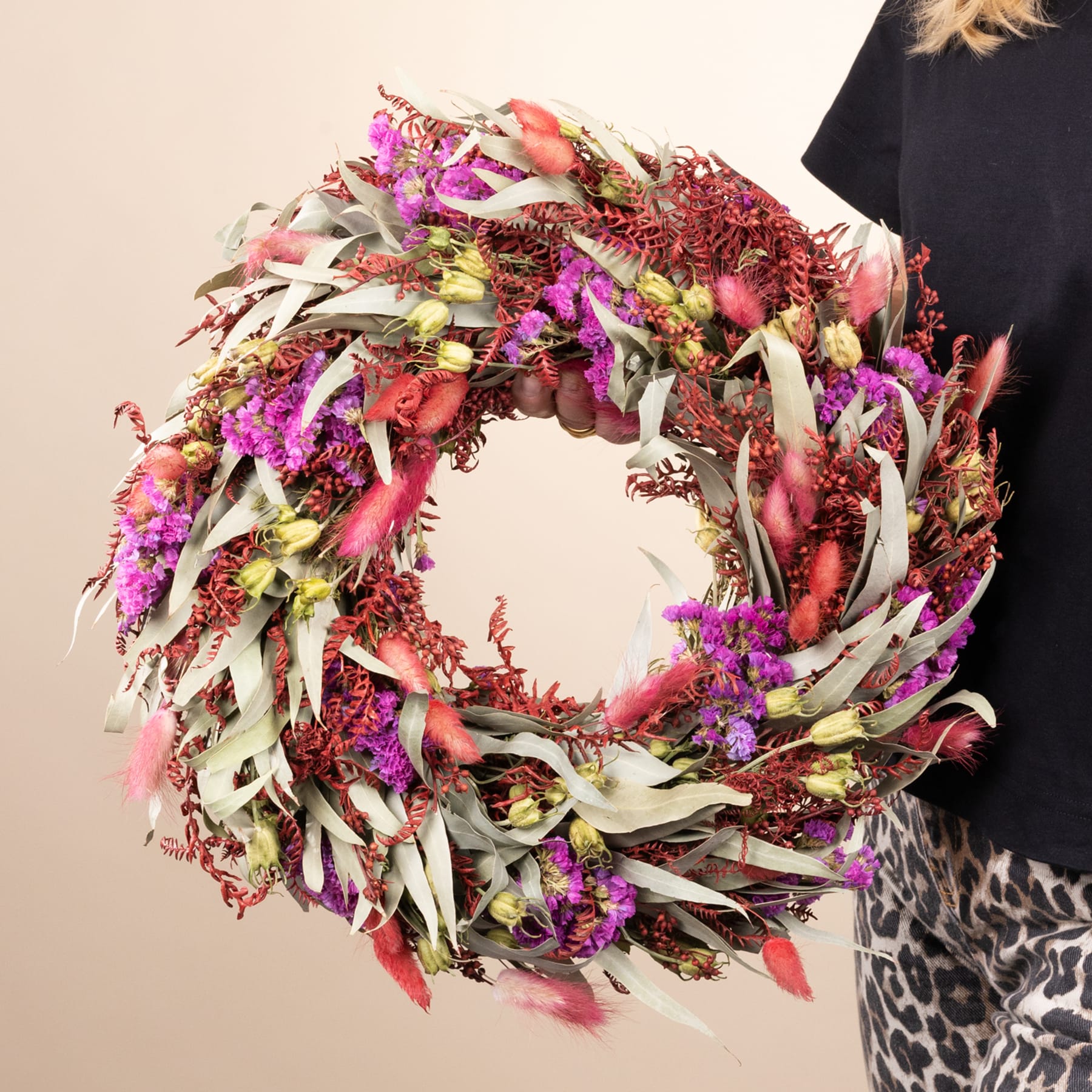 Ein leuchtend bunter Kranz aus verschiedenen getrockneten Blumen und Blättern wird von einer Person gehalten, die ein schwarz-weiß gemustertes Kleidungsstück trägt.