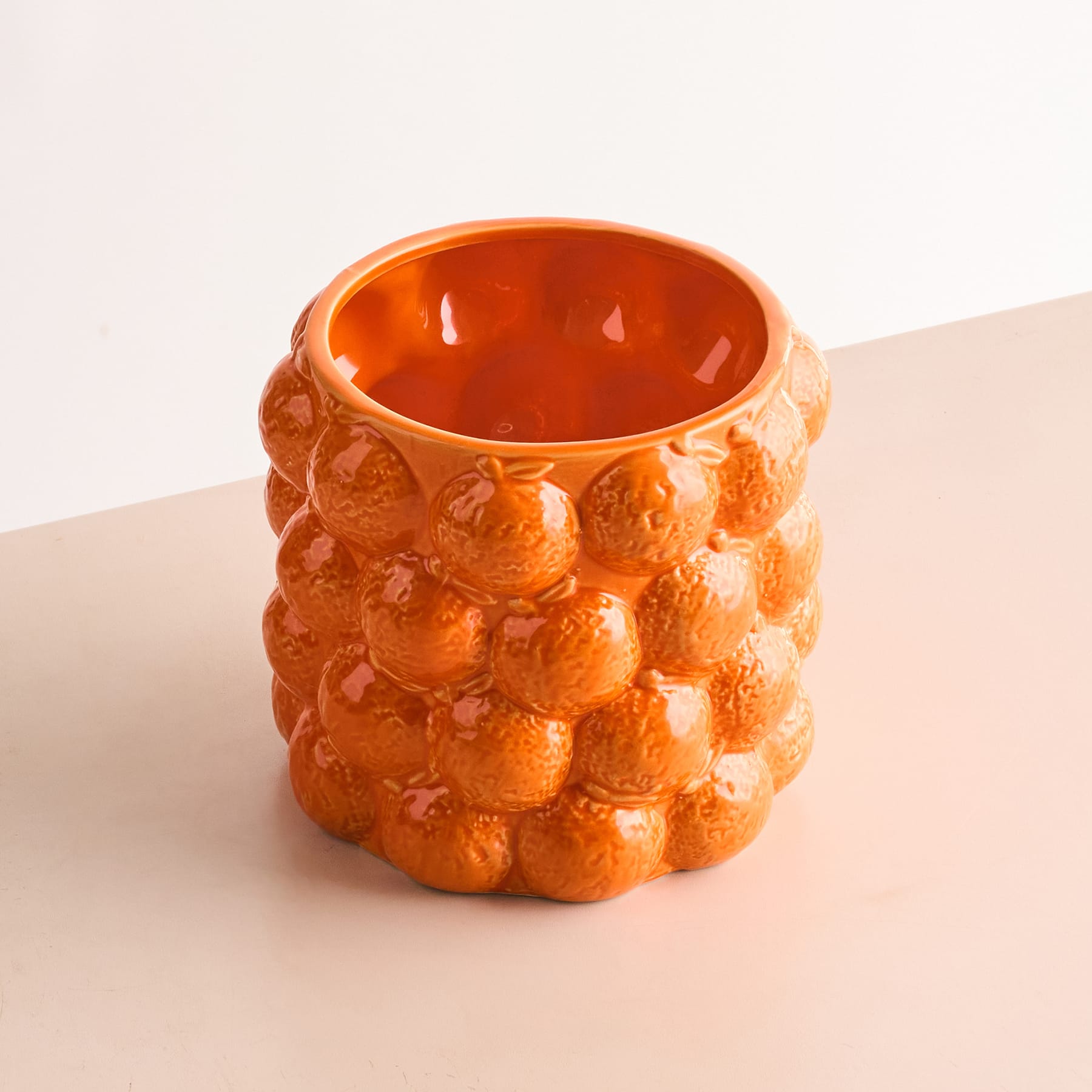 Eine leuchtend orangefarbene Keramikvase oder ein Keramikbehälter mit einer strukturierten, ananasähnlichen Oberfläche steht auf einer hellen Oberfläche vor einem schlichten Hintergrund.