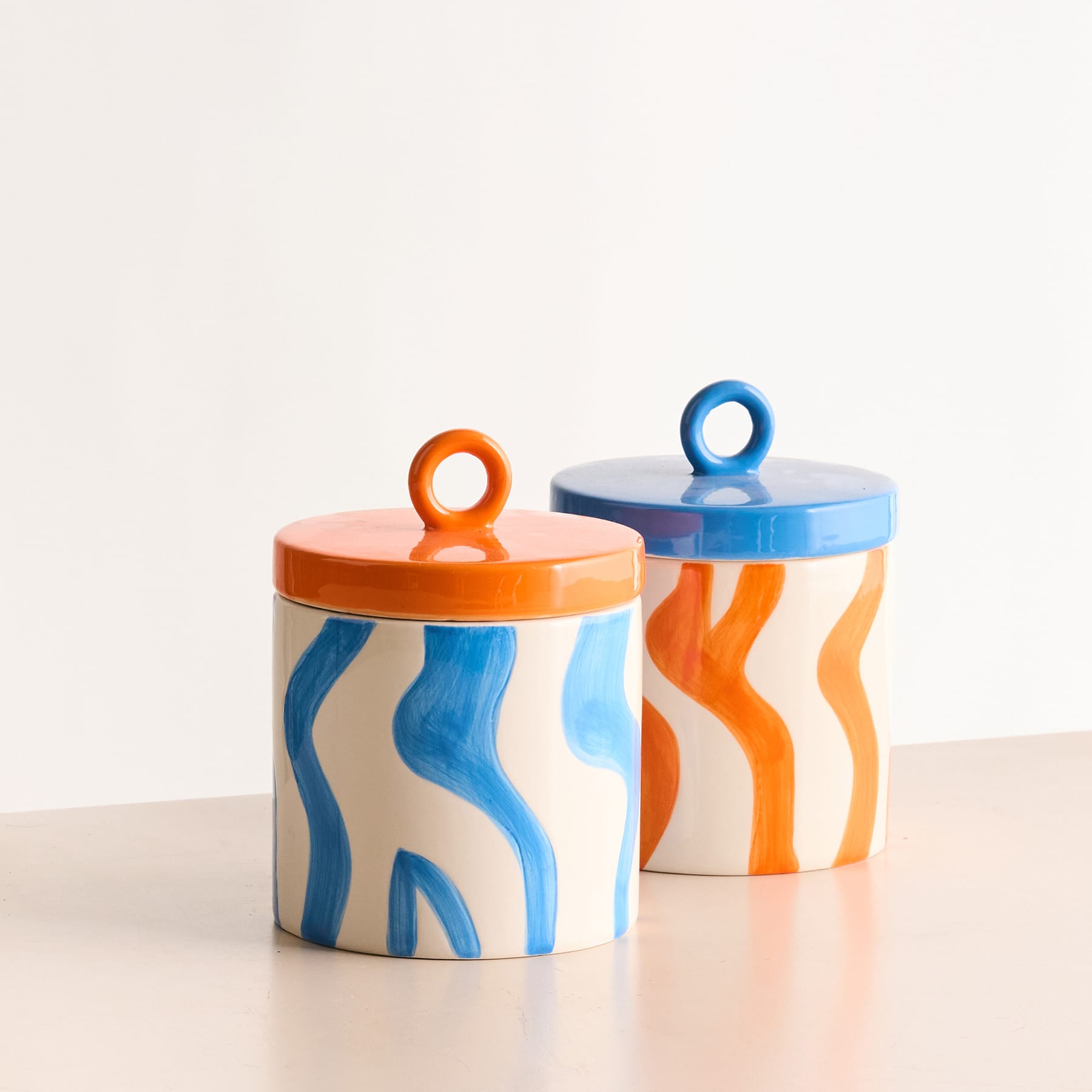 Zwei bunte Keramikgefäße mit abstrakten Wellenmustern in Blau- und Orangetönen stehen auf einem schlichten weißen Hintergrund.