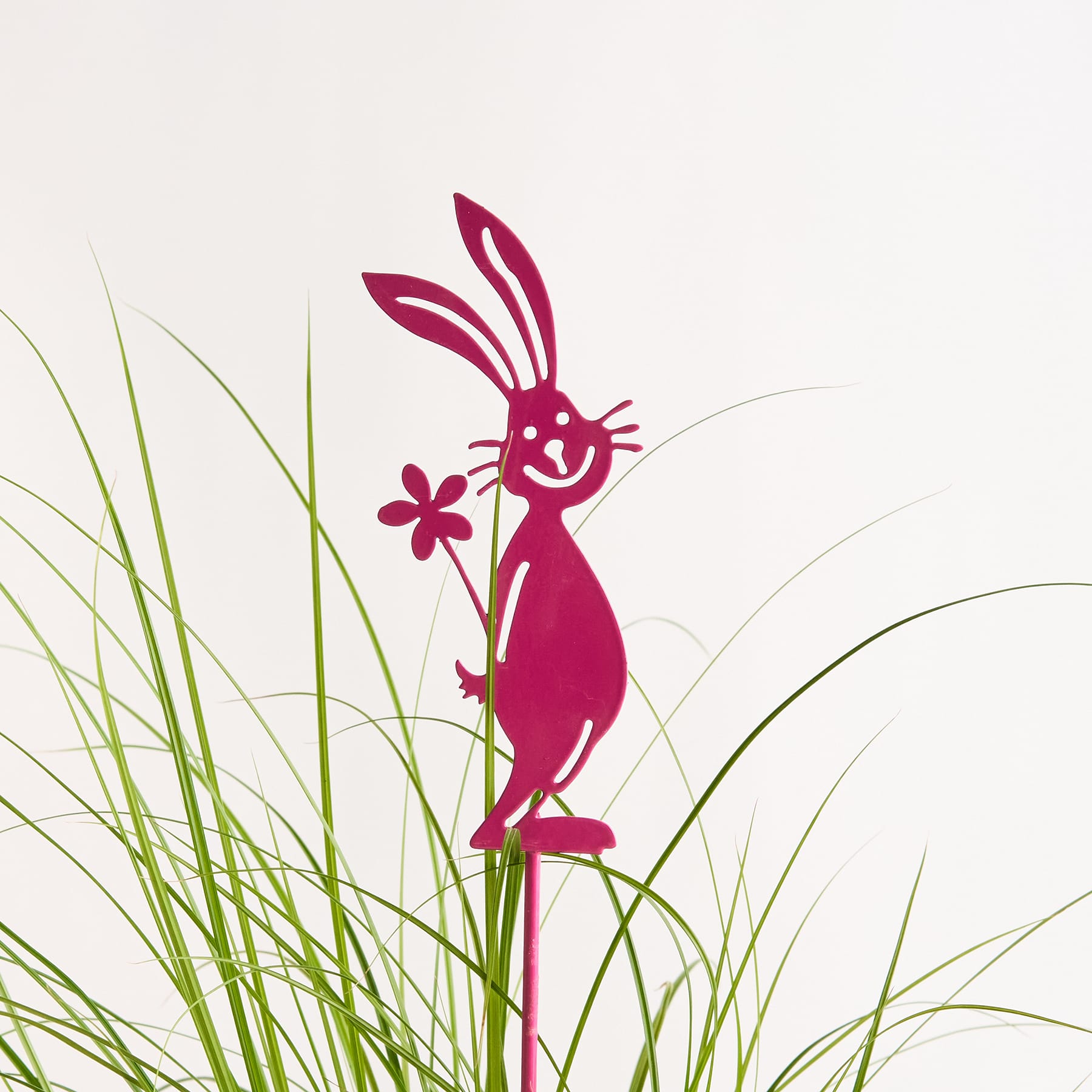 Eine leuchtend rosa, kaninchenähnliche Figur steht inmitten üppigen grünen Grases und hält eine kleine Blume in ihrer Pfote.