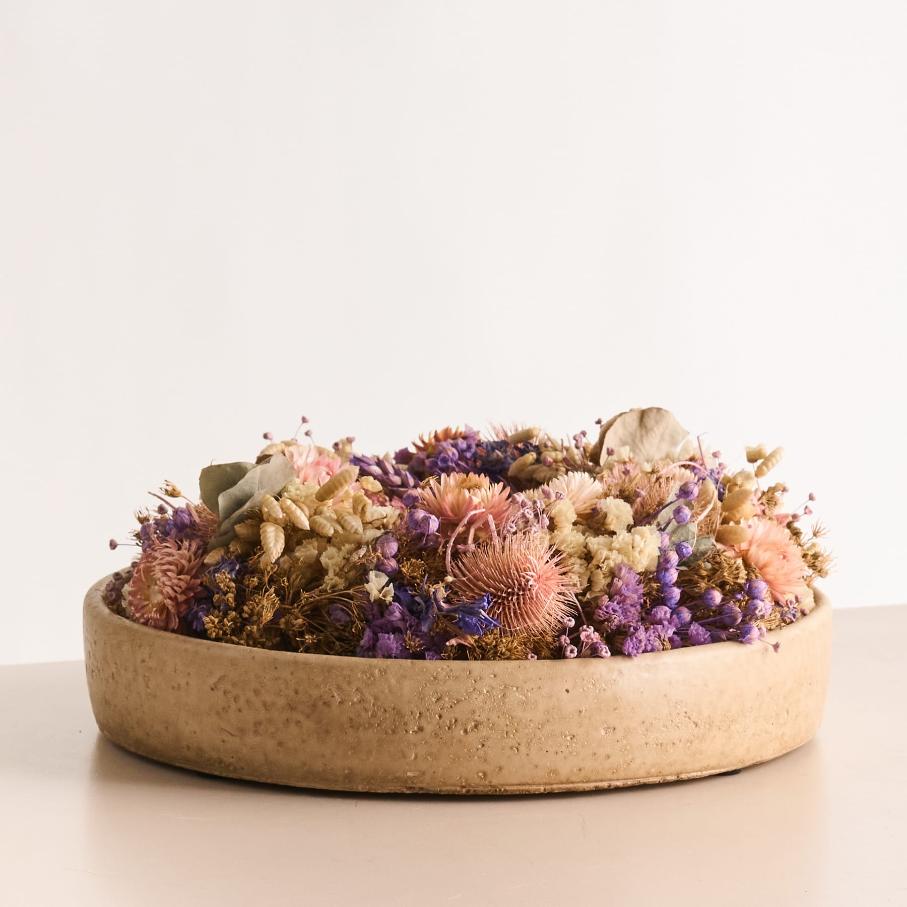 Auf einem schlichten weißen Hintergrund steht eine Keramikschale mit einem farbenfrohen Arrangement getrockneter Blumen in Lila-, Gelb- und Rosatönen.