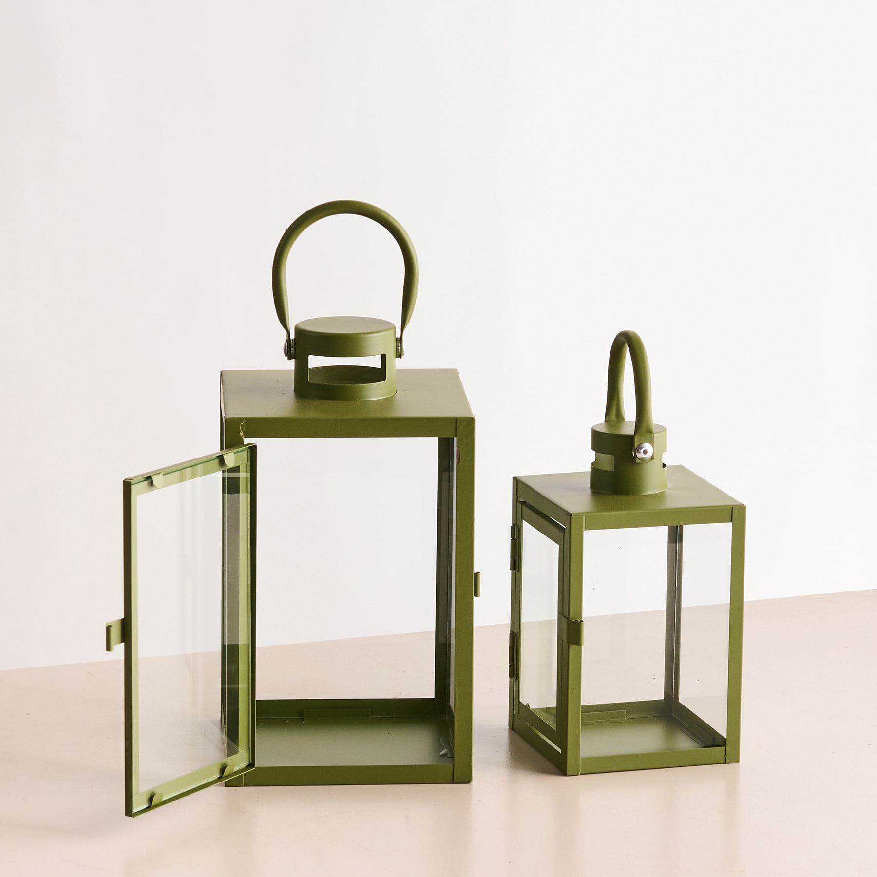 Zwei olivgrüne Metalllaternen mit Glasplatten stehen auf einer hellen Oberfläche und schaffen eine minimalistische und moderne Ästhetik.