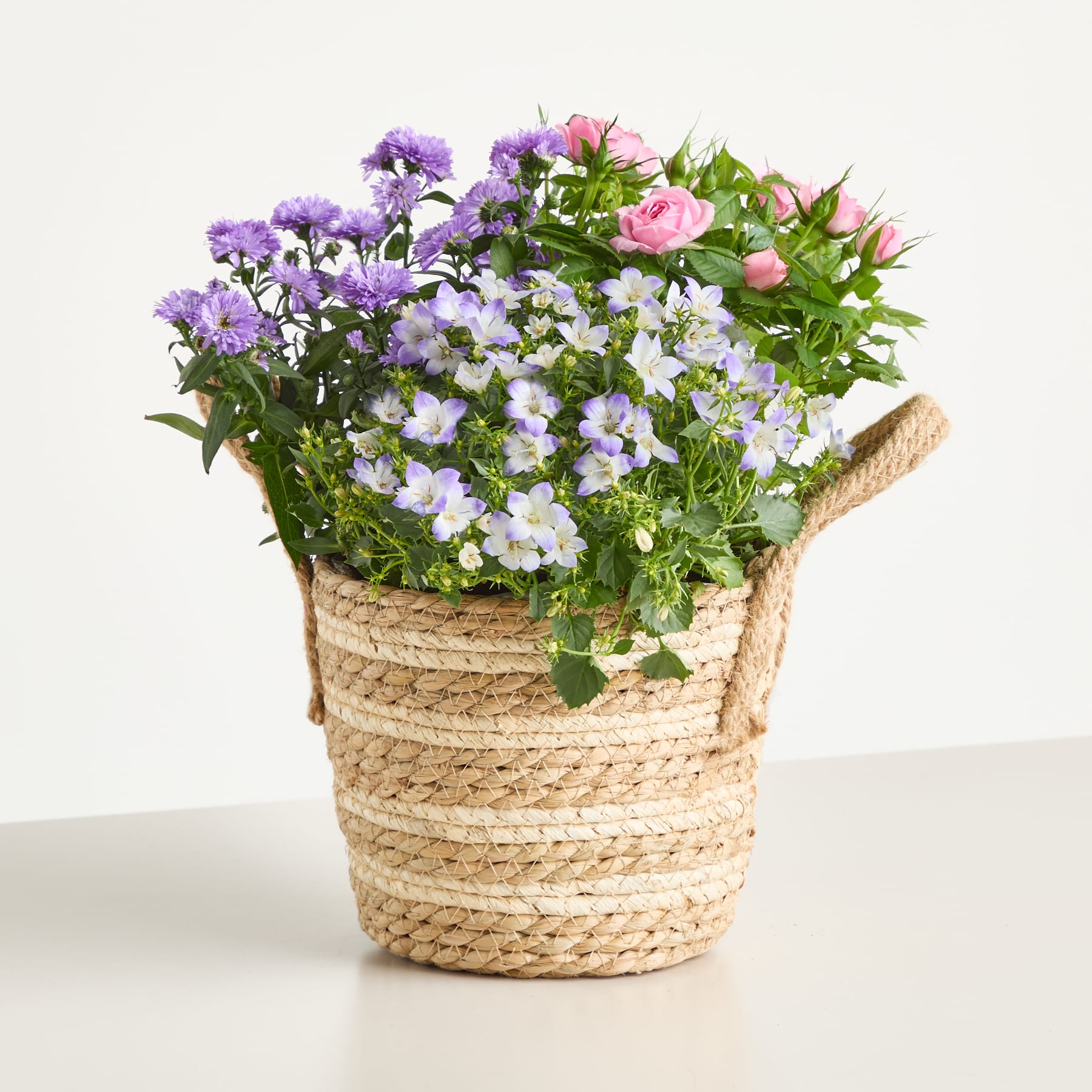Ein geflochtener Korb, gefüllt mit einem farbenfrohen Arrangement aus violetten, rosa und weißen Blumen vor einem schlichten weißen Hintergrund. (Beschreibungstext KI-generiert)
