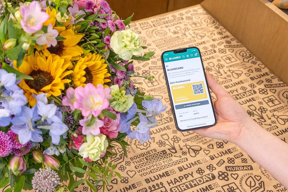 Das Bild zeigt eine Hand, die ein Smartphone mit der Benutzeroberfläche einer mobilen App hält, umgeben von einem farbenfrohen Strauß bunter Blumen, darunter Sonnenblumen, Gänseblümchen und andere Blüten.