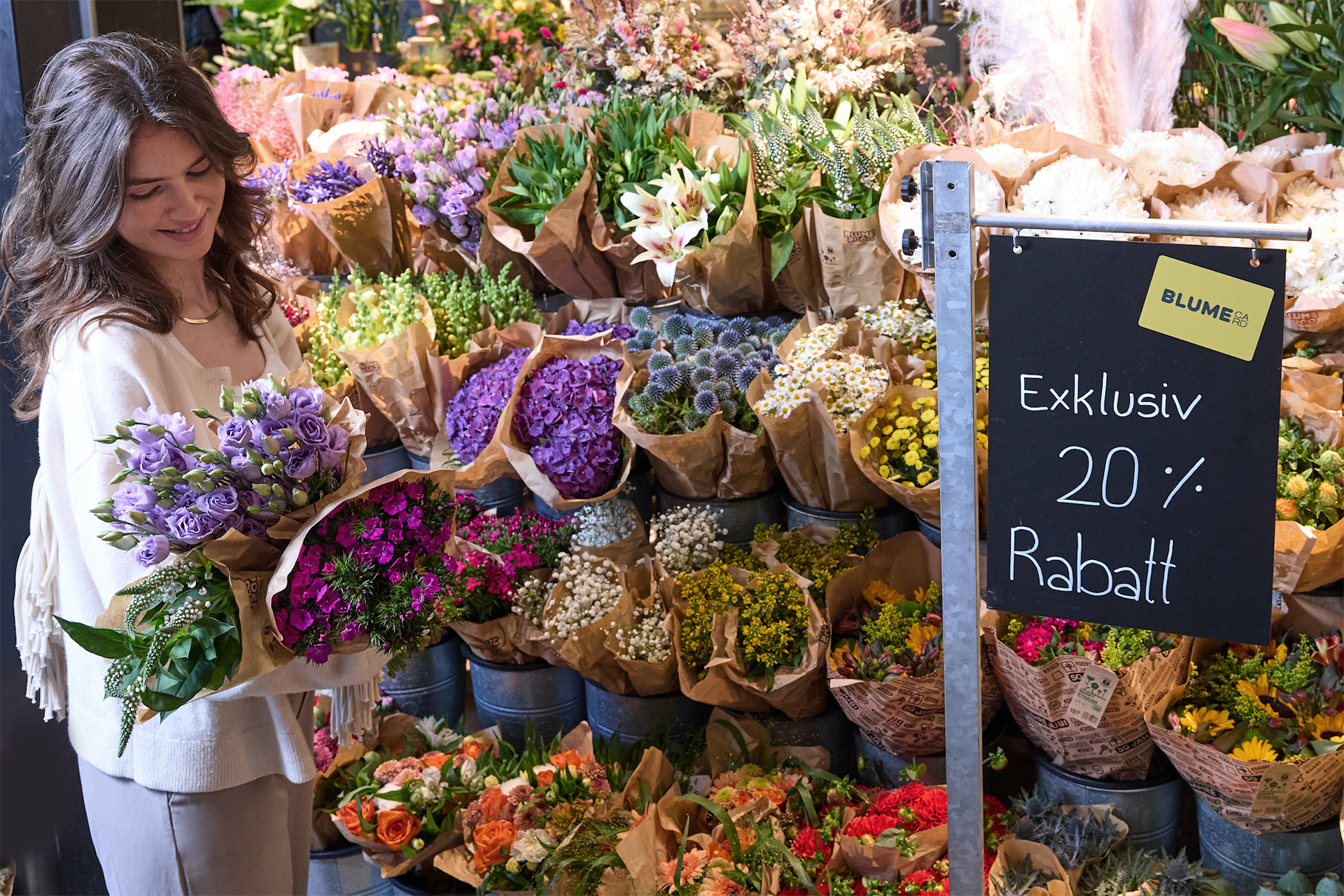 Eine Frau steht in einem lebhaften Blumenladen, umgeben von einer Reihe bunter Blumensträuße und einem Schild, das für einen Rabatt von 20 % wirbt.