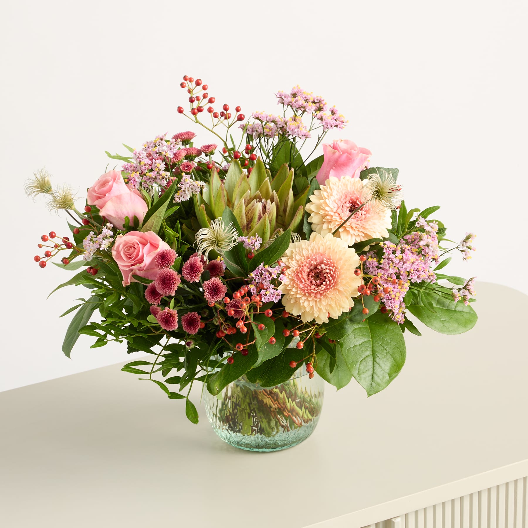 Ein farbenfroher Strauß verschiedener rosa und weißer Blumen, darunter Rosen, Dahlien und andere Blüten, arrangiert in einer Glasvase vor einem schlichten weißen Hintergrund.