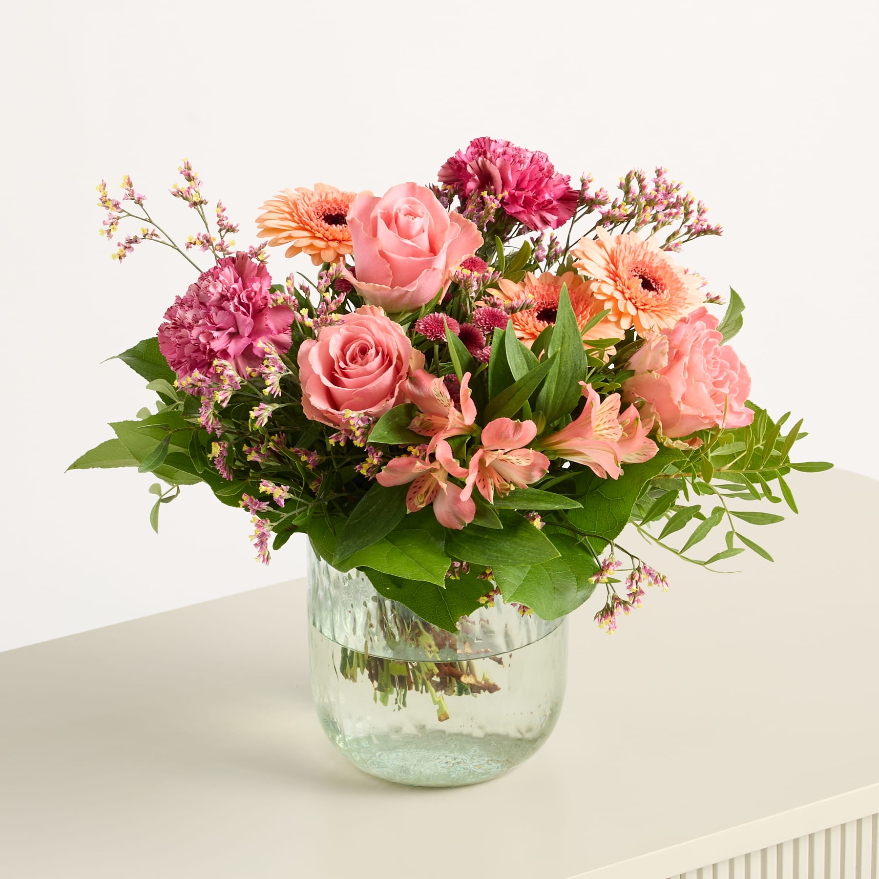 Ein leuchtender Strauß rosa und orangefarbener Blumen, darunter Rosen, Gerbera und Nelken, arrangiert in einer Glasvase vor einem schlichten weißen Hintergrund.