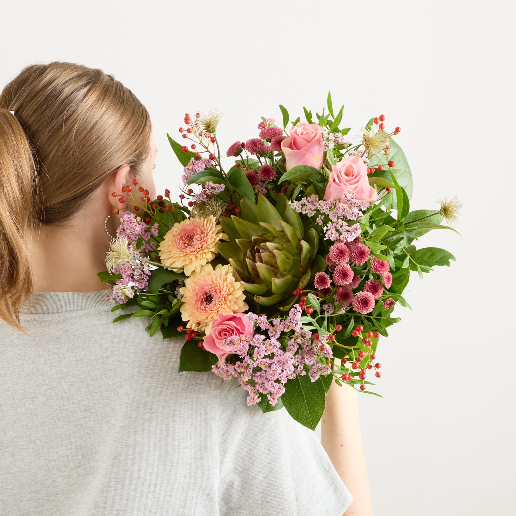 Im Vordergrund steht ein leuchtender Strauß bunter Blumen, darunter rosa Rosen, gelbe Gänseblümchen und eine große Artischocke, während der Hintergrund eine schlichte weiße Fläche ist.