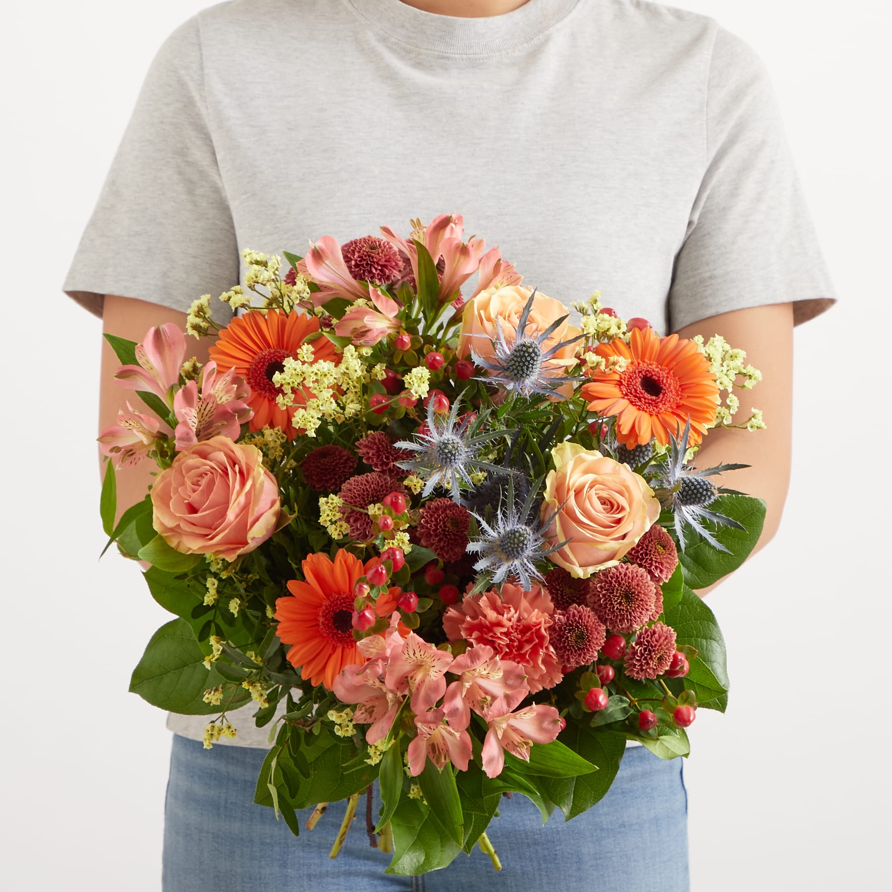 Ein lebendiger Strauß bunter Blumen, darunter Rosen, Gerbera und Nelken, steht im Vordergrund vor einem schlichten weißen Hintergrund.