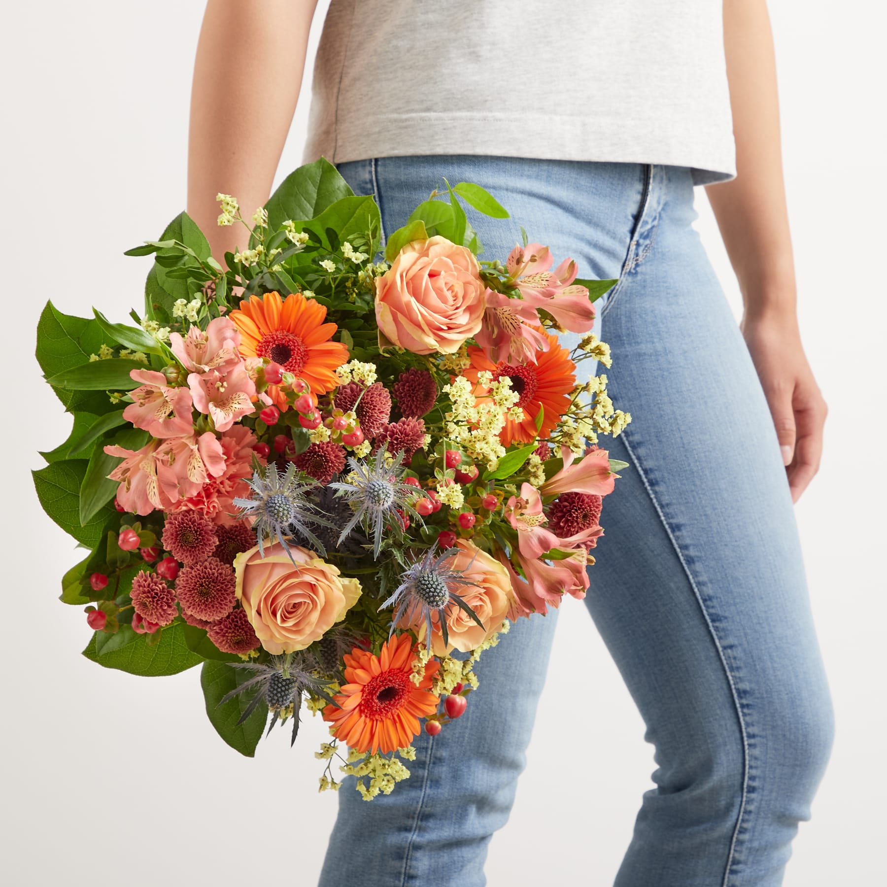 Ein leuchtender Strauß bunter Blumen, darunter Rosen, Gerbera und Grünpflanzen, wird vor eine Person in Jeans gehalten.