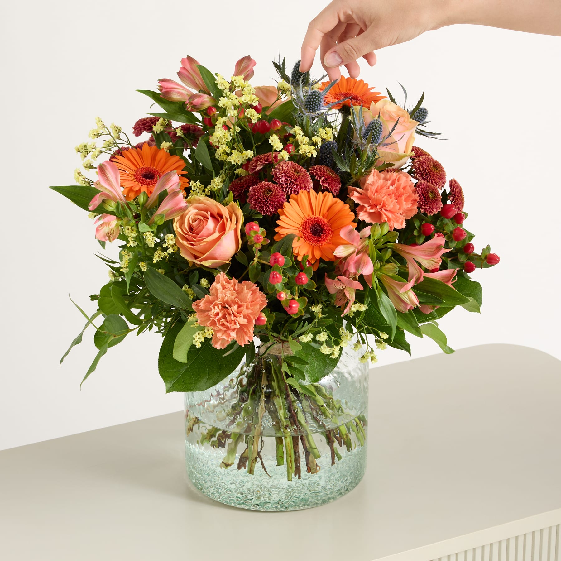 Ein lebendiger Strauß bunter Blumen, darunter Rosen, Gerbera und Nelken, arrangiert in einer Glasvase vor einem schlichten weißen Hintergrund.