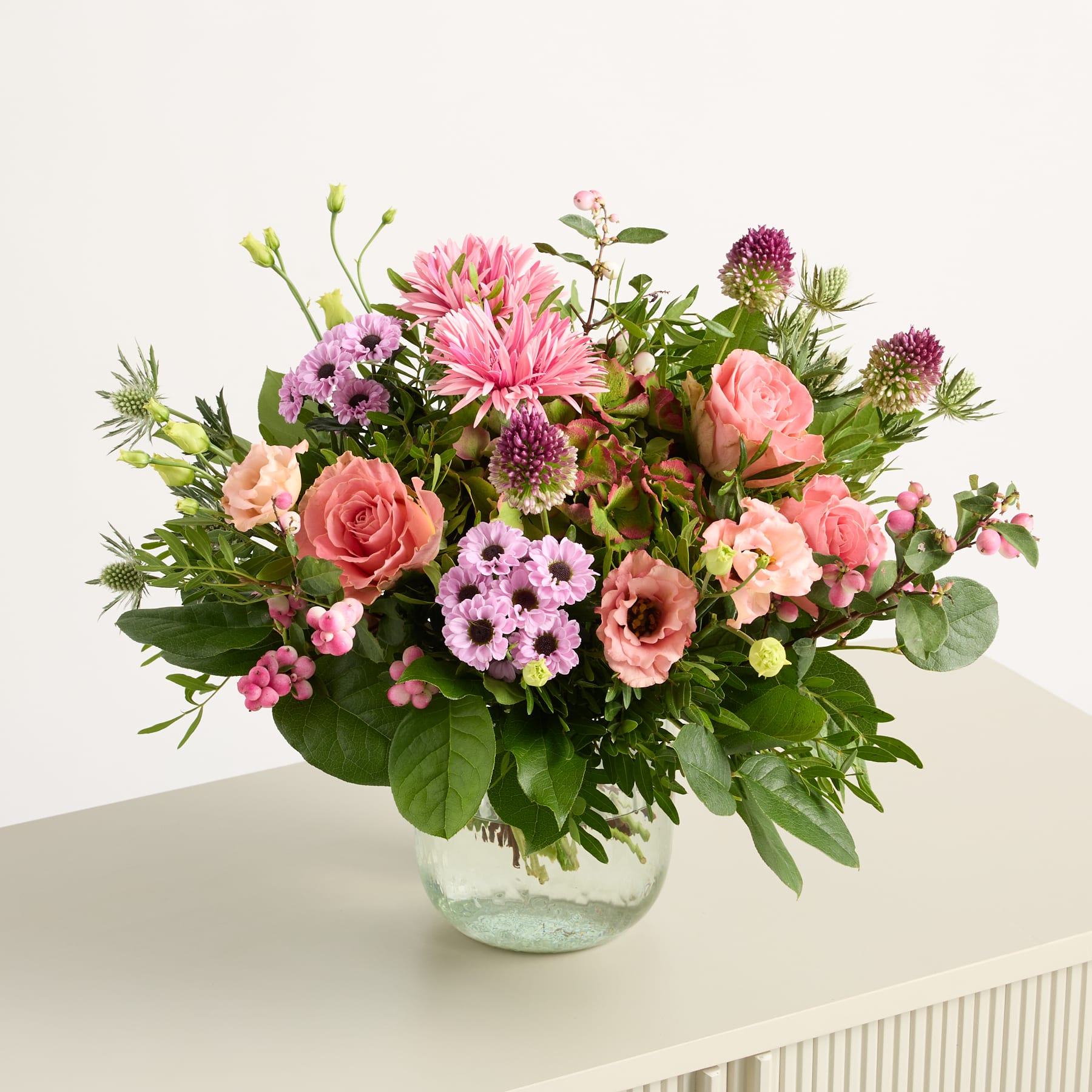 Ein farbenfroher Strauß verschiedener rosa und violetter Blumen, darunter Rosen, Dahlien und Chrysanthemen, arrangiert in einer Glasvase vor einem schlichten weißen Hintergrund.