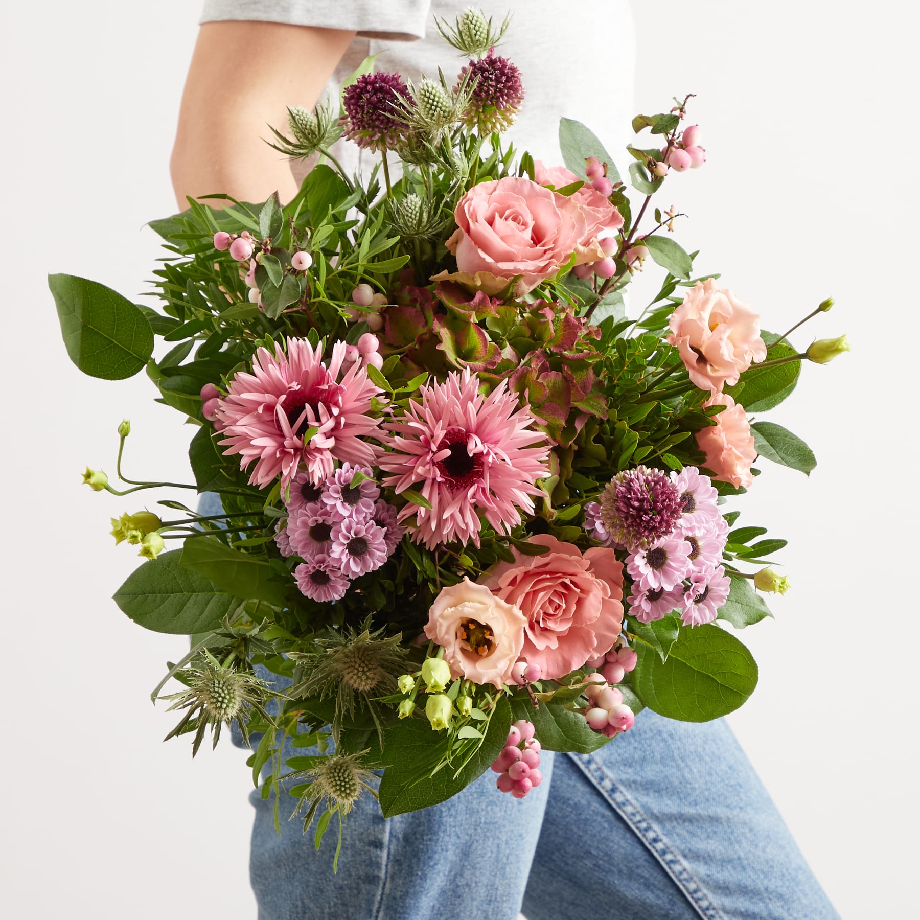 Ein farbenfroher Strauß rosa und violetter Blumen, darunter Rosen, Gänseblümchen und andere Blüten, wird vor die Beine einer Person gehalten, die Blue Jeans trägt.