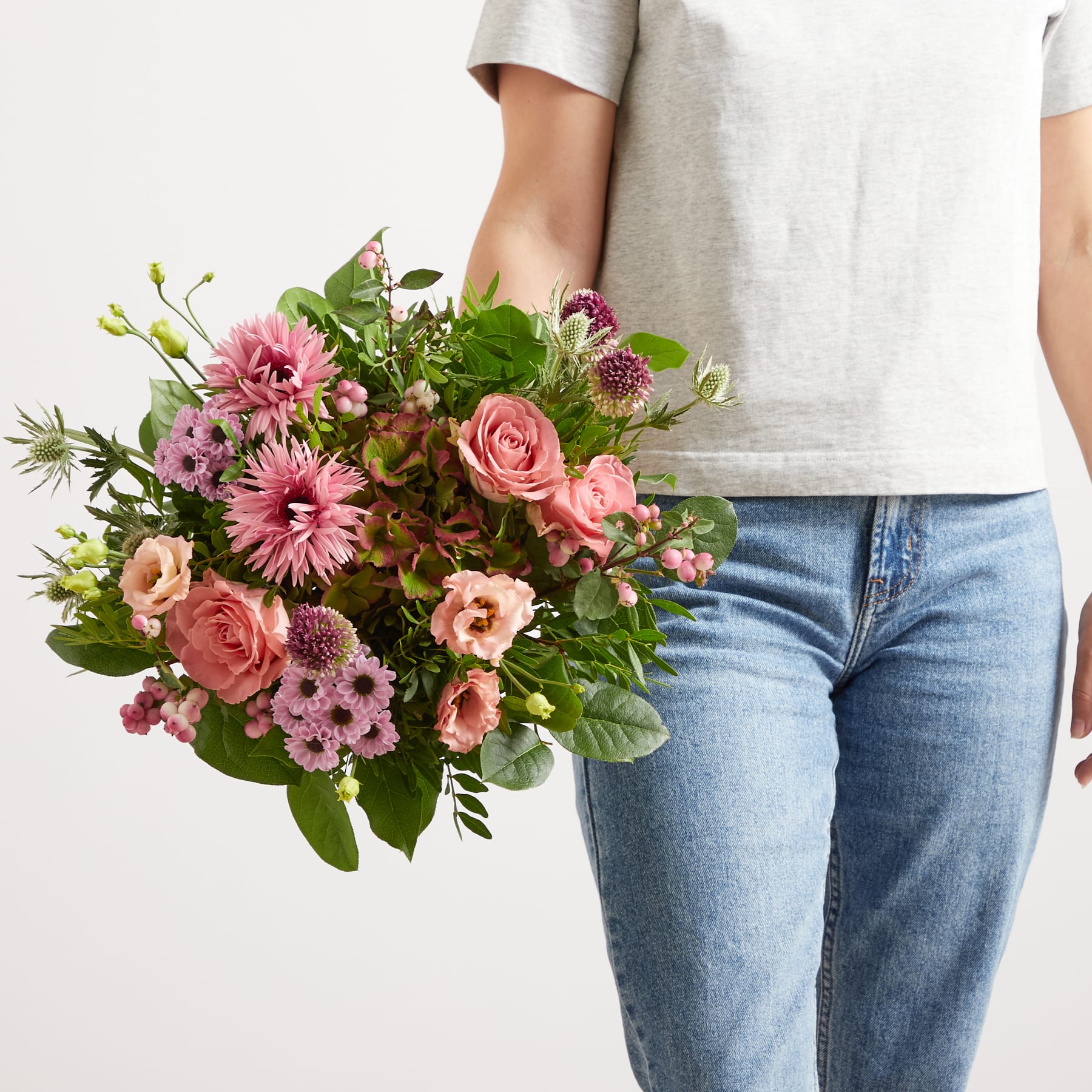 Ein farbenfroher Strauß rosa und weißer Blumen, darunter Rosen und Gänseblümchen, wird vor einer Person gehalten, die ein helles Oberteil und Blue Jeans vor einem schlichten weißen Hintergrund trägt.