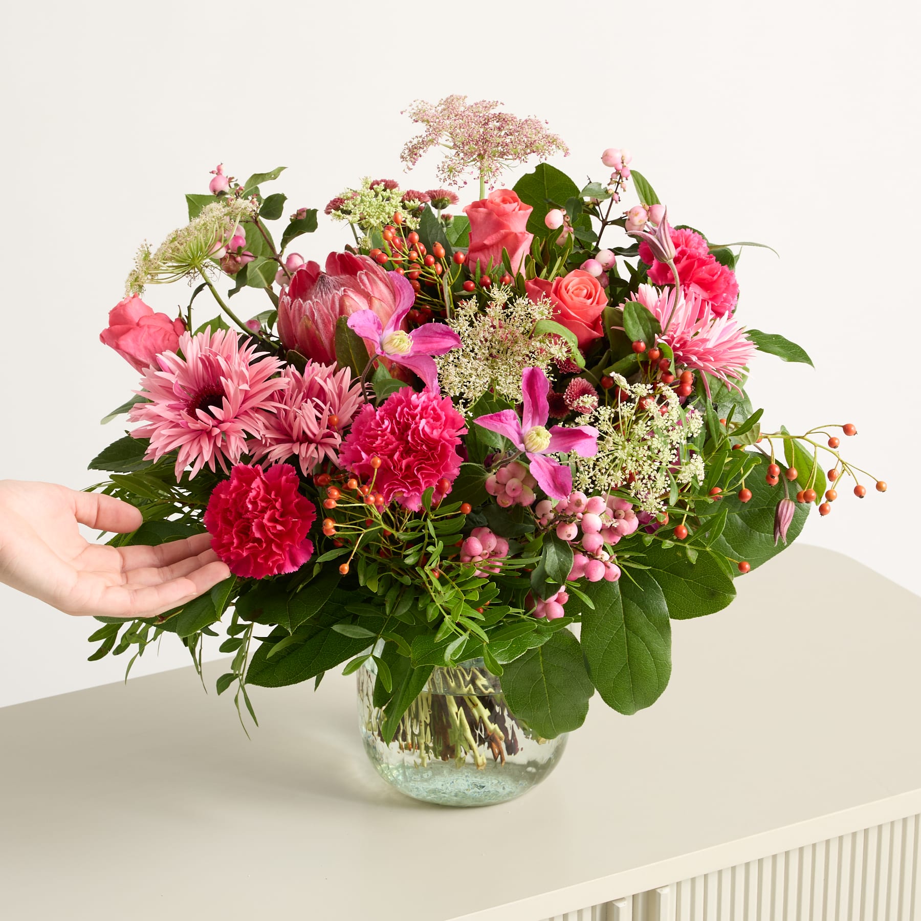 Ein farbenfroher Strauß verschiedener rosa und roter Blumen, darunter Nelken, Rosen und Gänseblümchen, steht in einer Glasvase vor einem schlichten weißen Hintergrund.