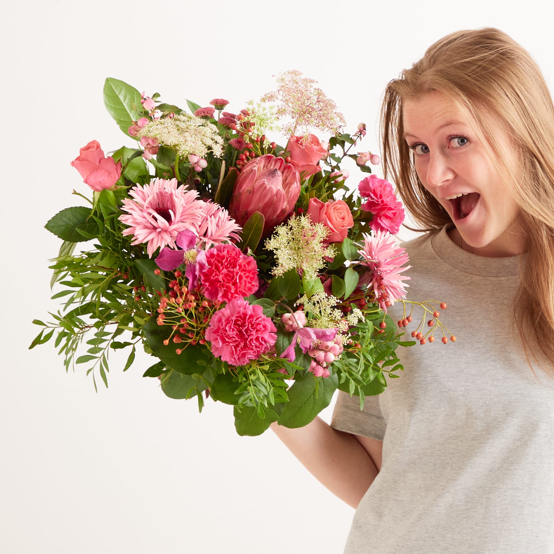 Eine junge Frau mit einem strahlenden, fröhlichen Gesichtsausdruck hält einen großen, leuchtenden Strauß bunter Blumen, darunter rosa Nelken, rote Rosen und weiße Gänseblümchen, vor einem schlichten weißen Hintergrund.