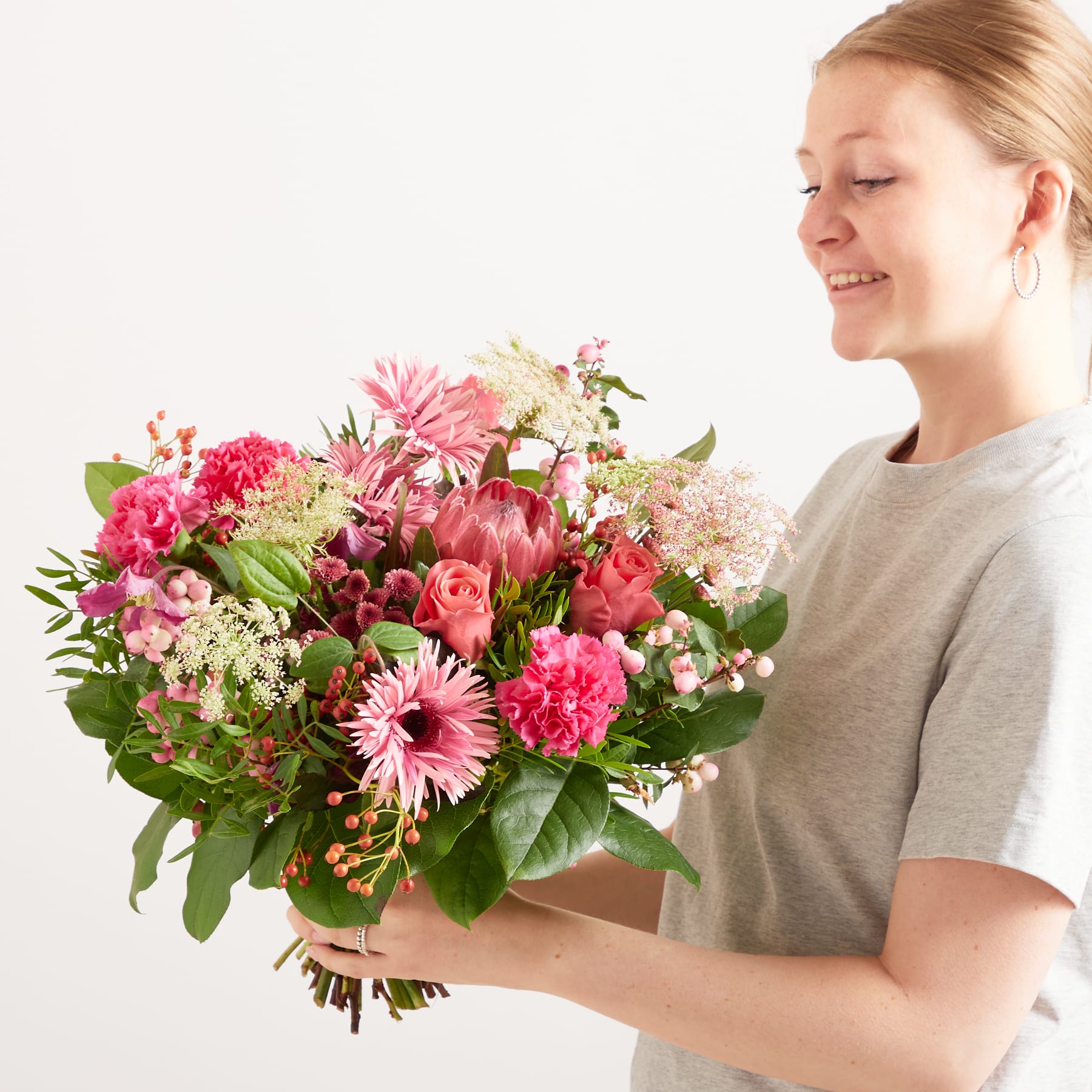 Eine junge Frau hält vor einem schlichten weißen Hintergrund einen farbenfrohen Strauß rosa und weißer Blumen, darunter Nelken und Gänseblümchen.