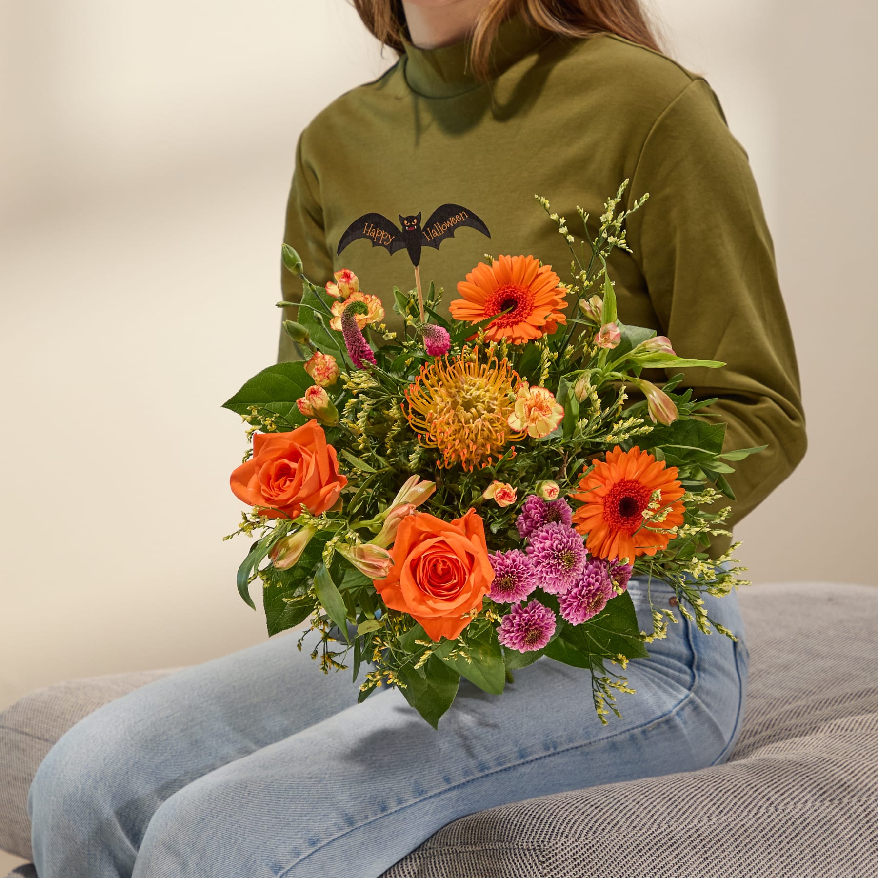 Ein farbenfroher Strauß orangefarbener und gelber Blumen, darunter Rosen, Chrysanthemen und eine Halloween-Dekoration in Fledermausform, vor einem schlichten weißen Hintergrund.