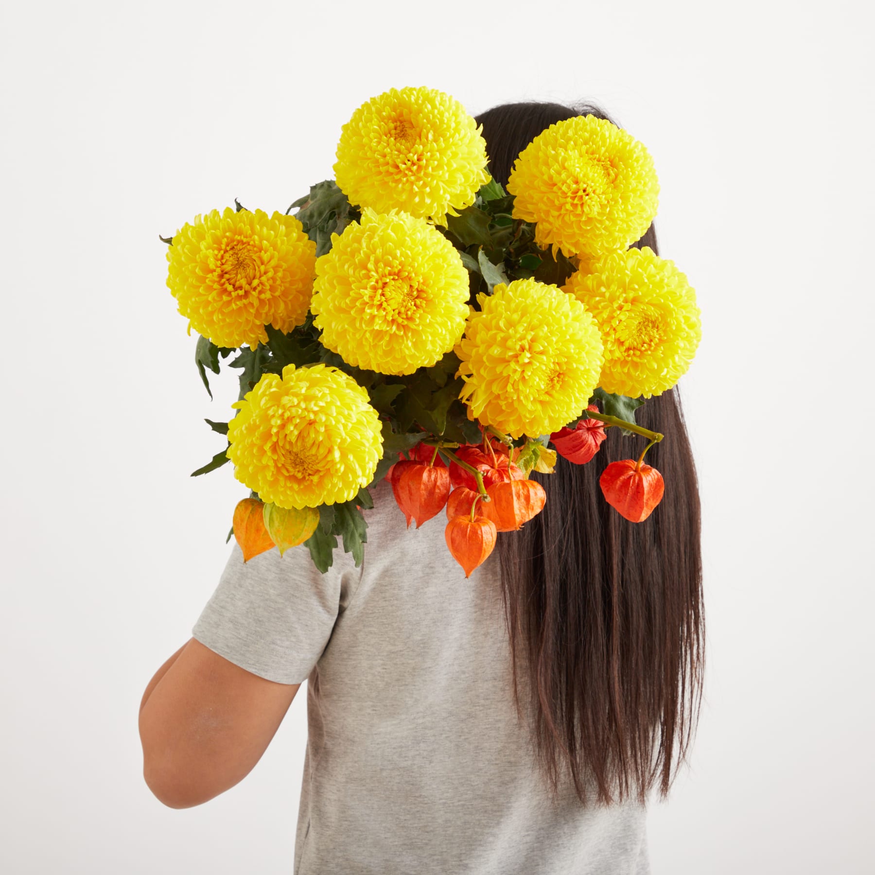 Ein Strauß leuchtend gelber Chrysanthemenblüten, gehalten von einer Person mit langem dunklem Haar vor einem schlichten weißen Hintergrund.