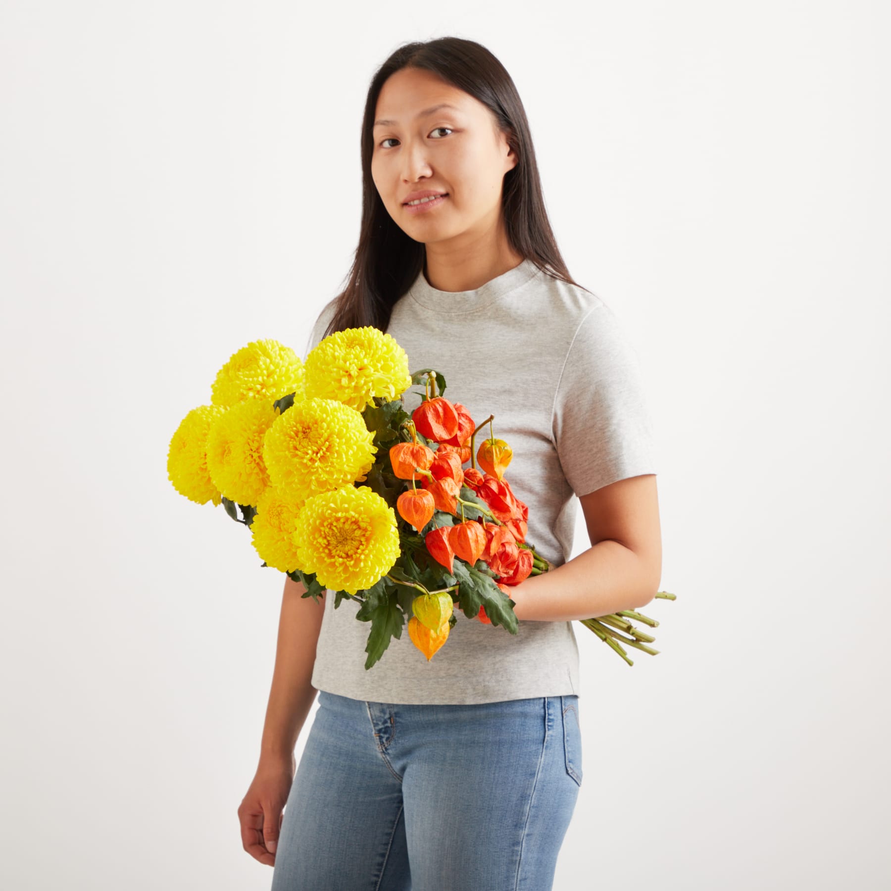 Eine junge Frau mit langem, dunklem Haar hält einen Strauß leuchtend gelber Chrysanthemen und roter Tulpen vor einem schlichten weißen Hintergrund.